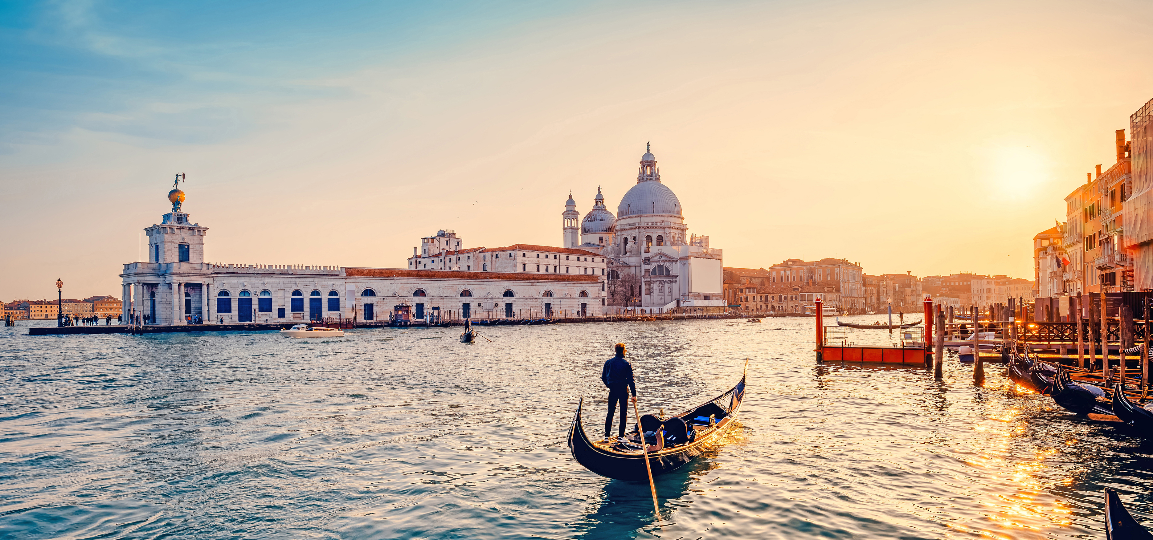 A gondola paddles along a river while the sun sits low in the sky.