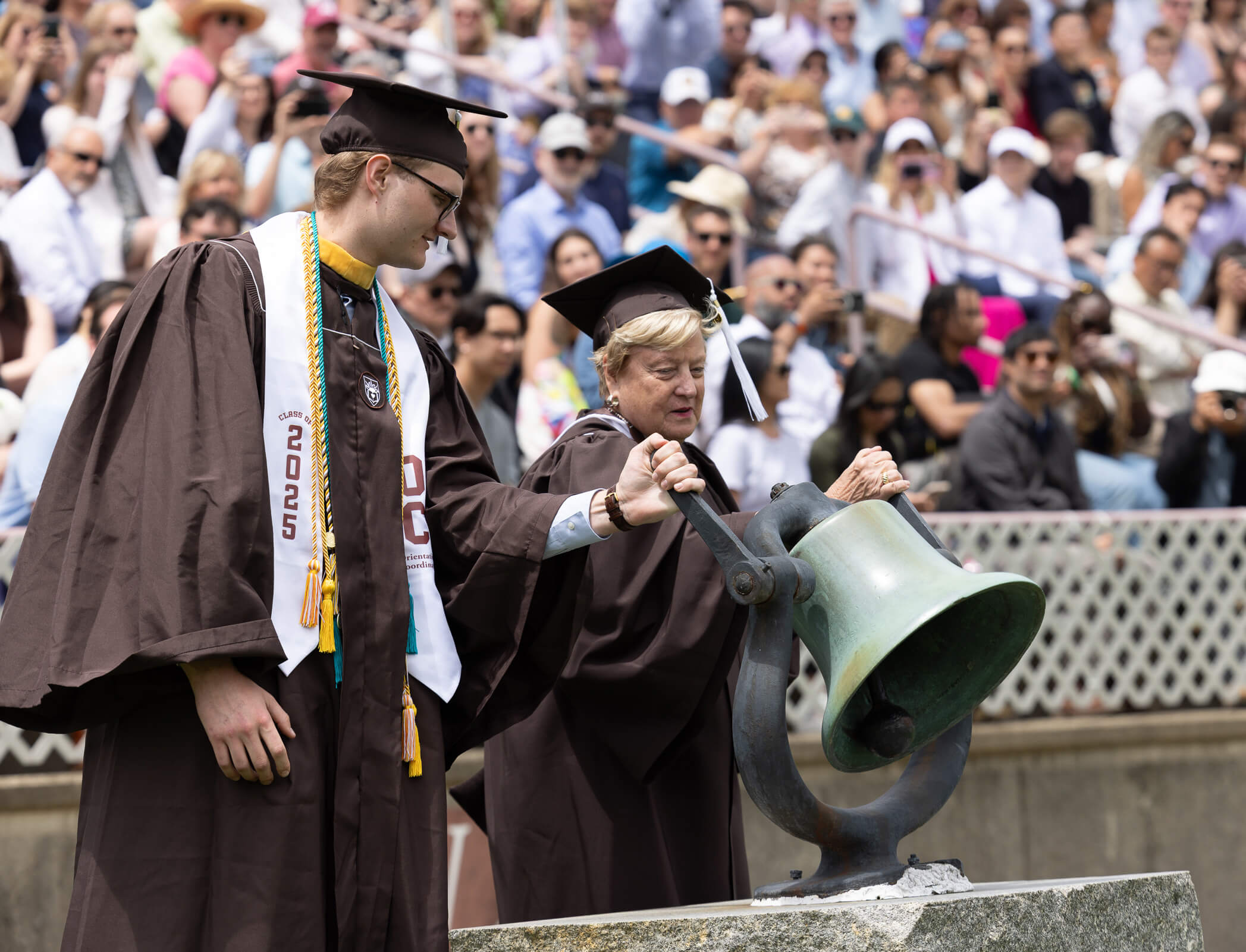 Both wearing brown graduation caps and gowns, a tall male student and an alumna from the class of 1975 both hold a large bell to ring it with a crowd in the stands behind them.