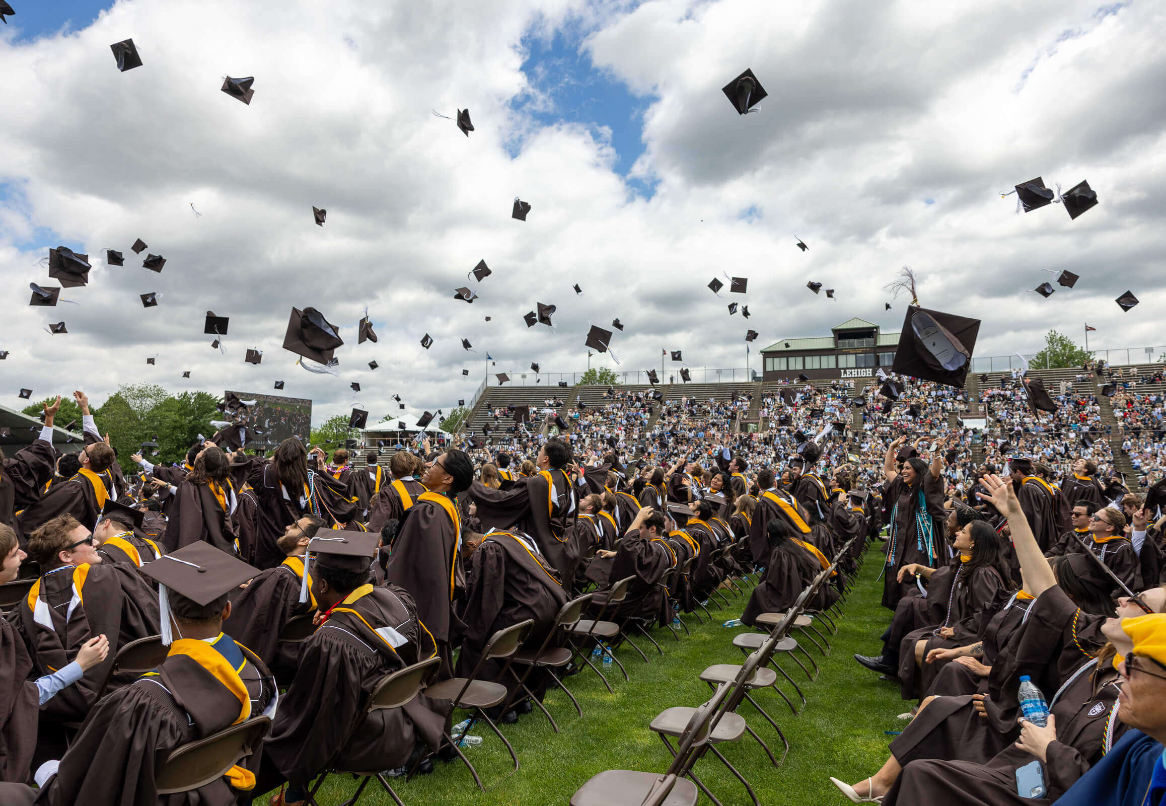 A wide shot shows a full crowd of graduates with caps tossed in the air with clouds visible in the blue sky.