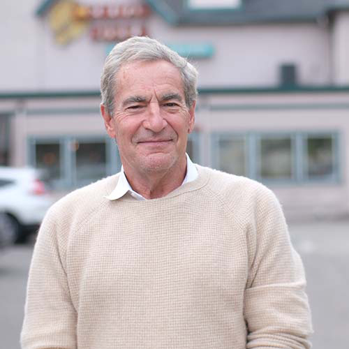 Craig wears a beige sweater and stands in front of a local pizza shop.