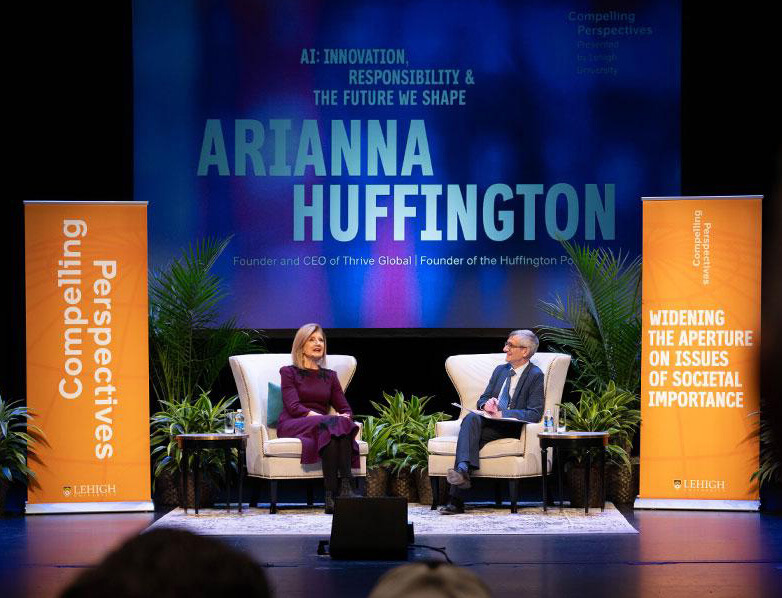 On a stage with a monitor behind them reading “Arianna Huffington”, Arianna Huffington sits with Joe Helble in large cream colored upholstered chairs.