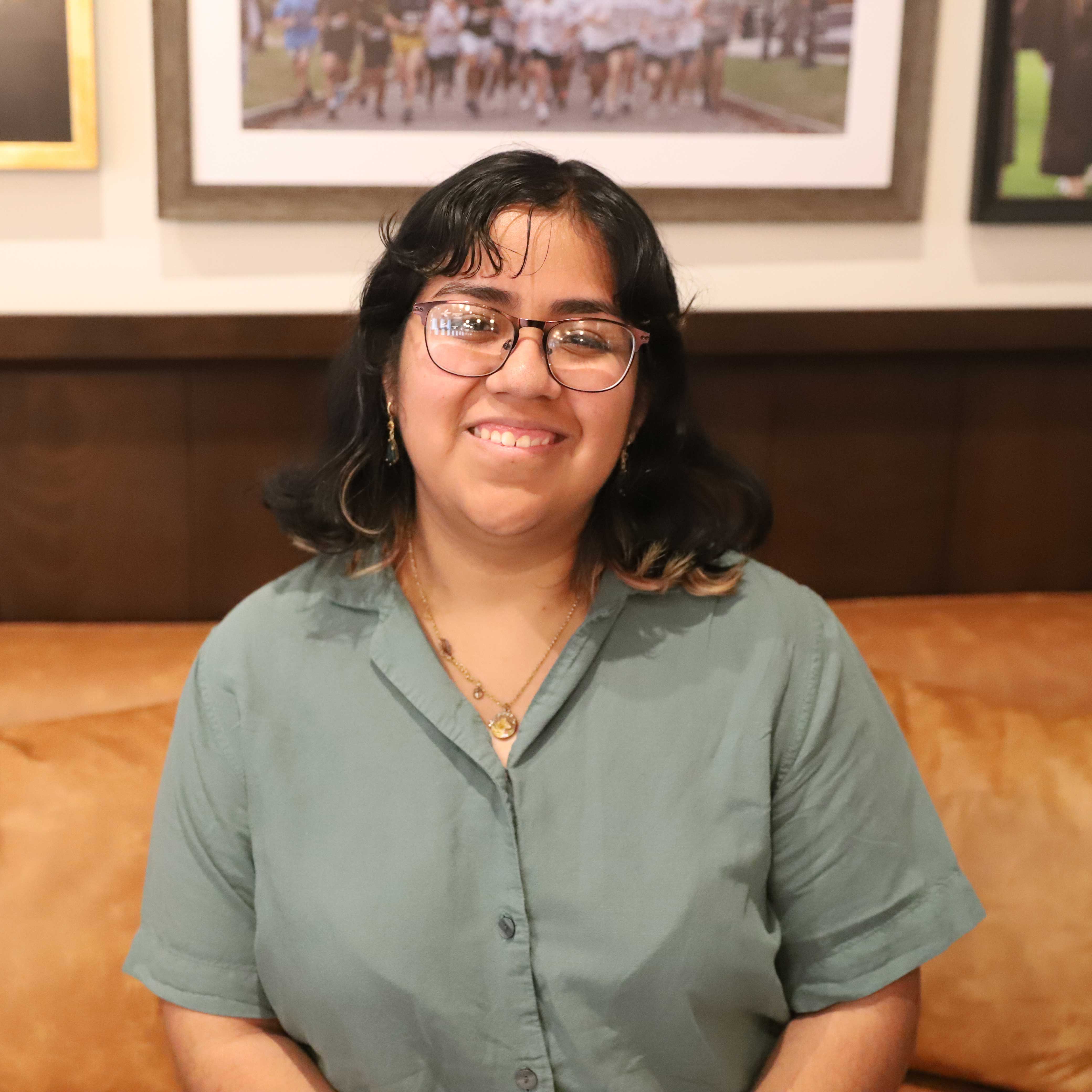 Crystal sits on a brown leather couch. She wears a sage button-down and glasses; photographs of students are behind her.