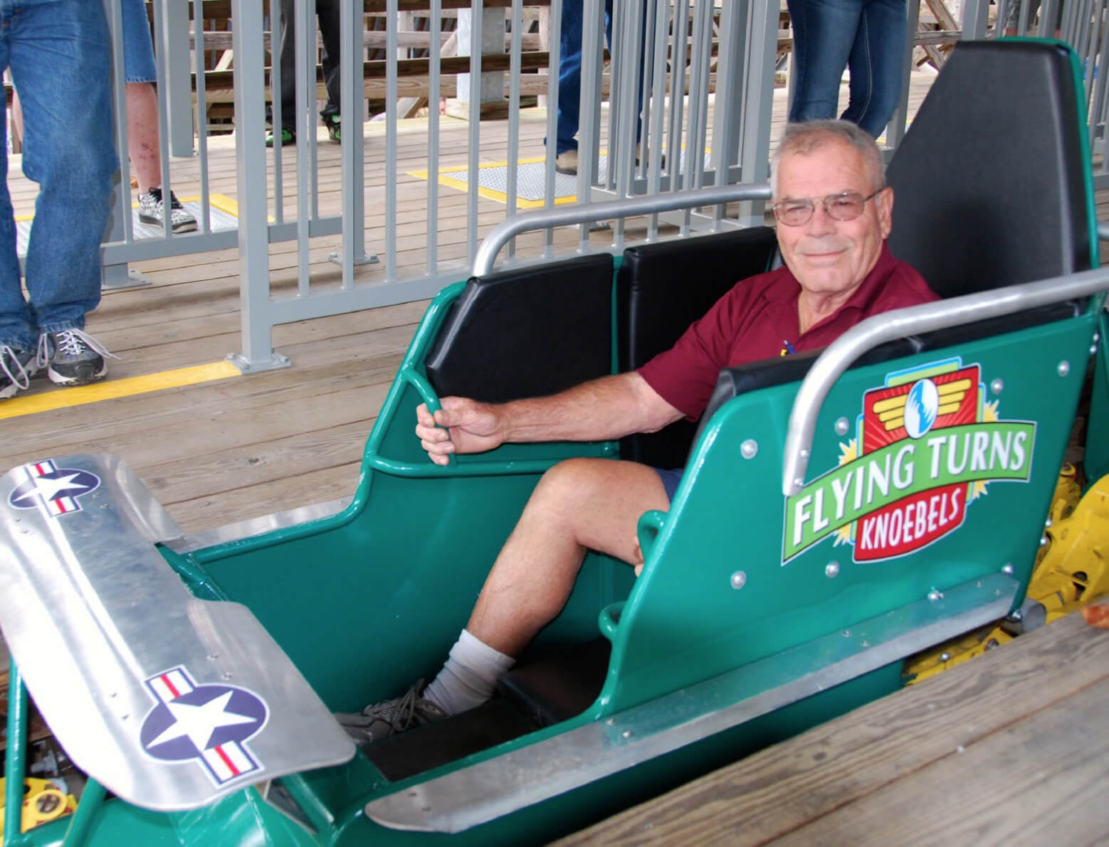 Dick Knoebel sits smiling in a green solo rider roller coaster cart that reads “Flying Turns- Knoebels” on the side.