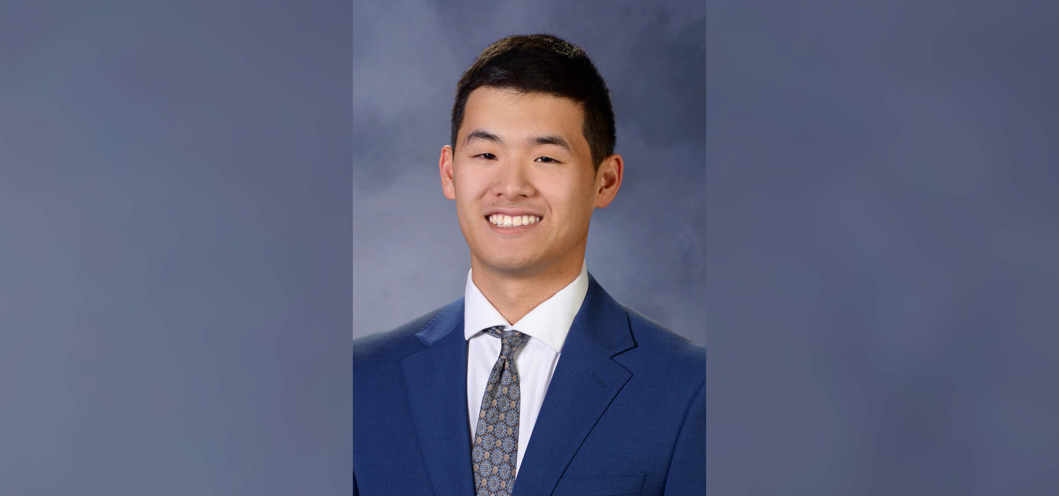 A man wears a navy suit, white shirt and patterned tie against a background with shades of dark blue and smiles for a headshot.