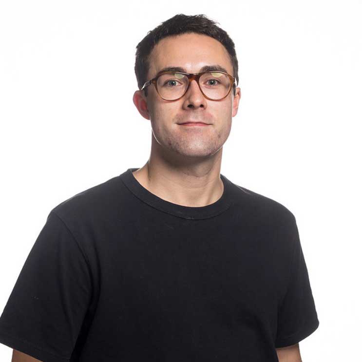 Daniel wears a black t-shirt and brown glasses. He stands in front of a white background and smiles.