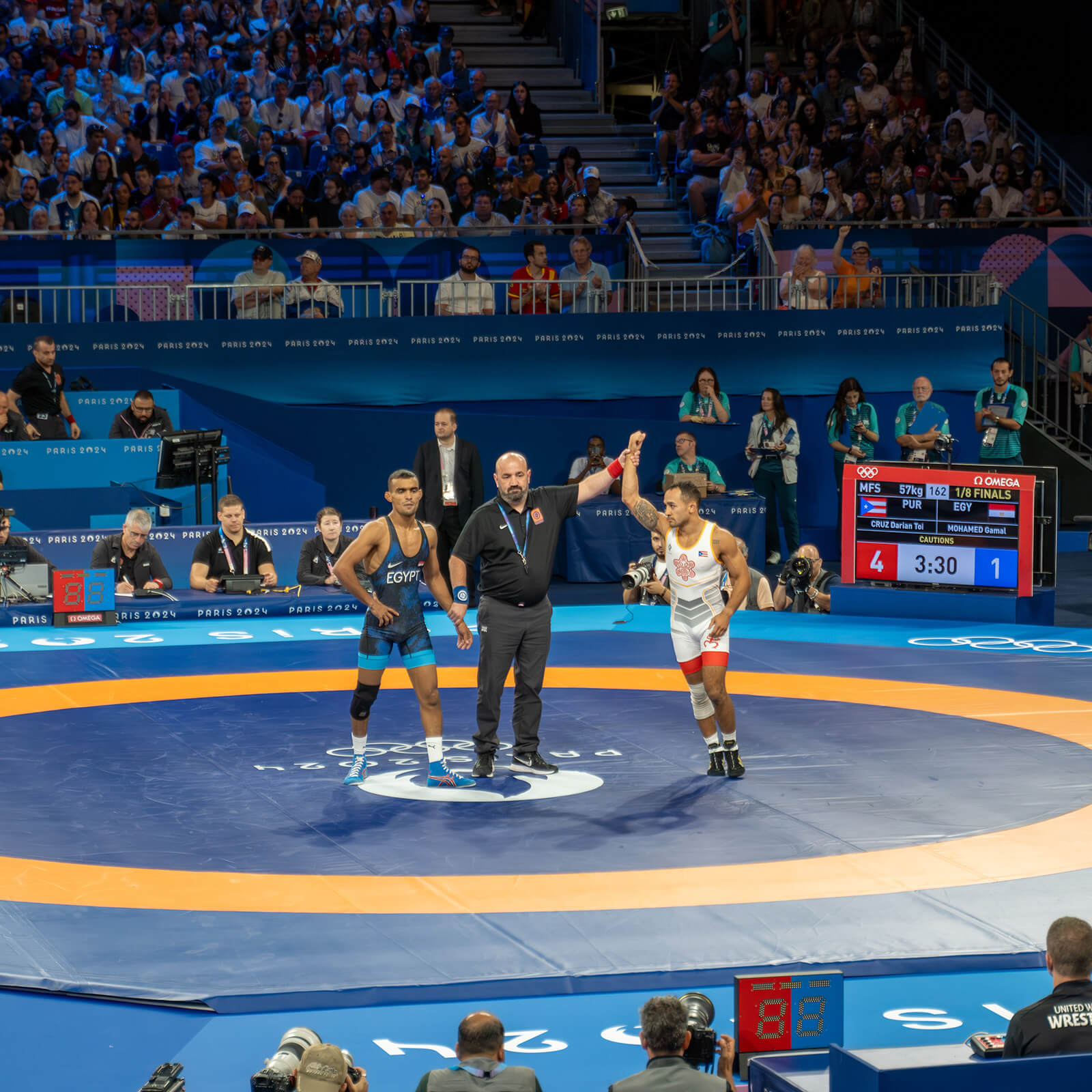 Referee stands in the center of the wrestling mat at the Olympics with two wrestlers, holding up Darian Cruz's hand.