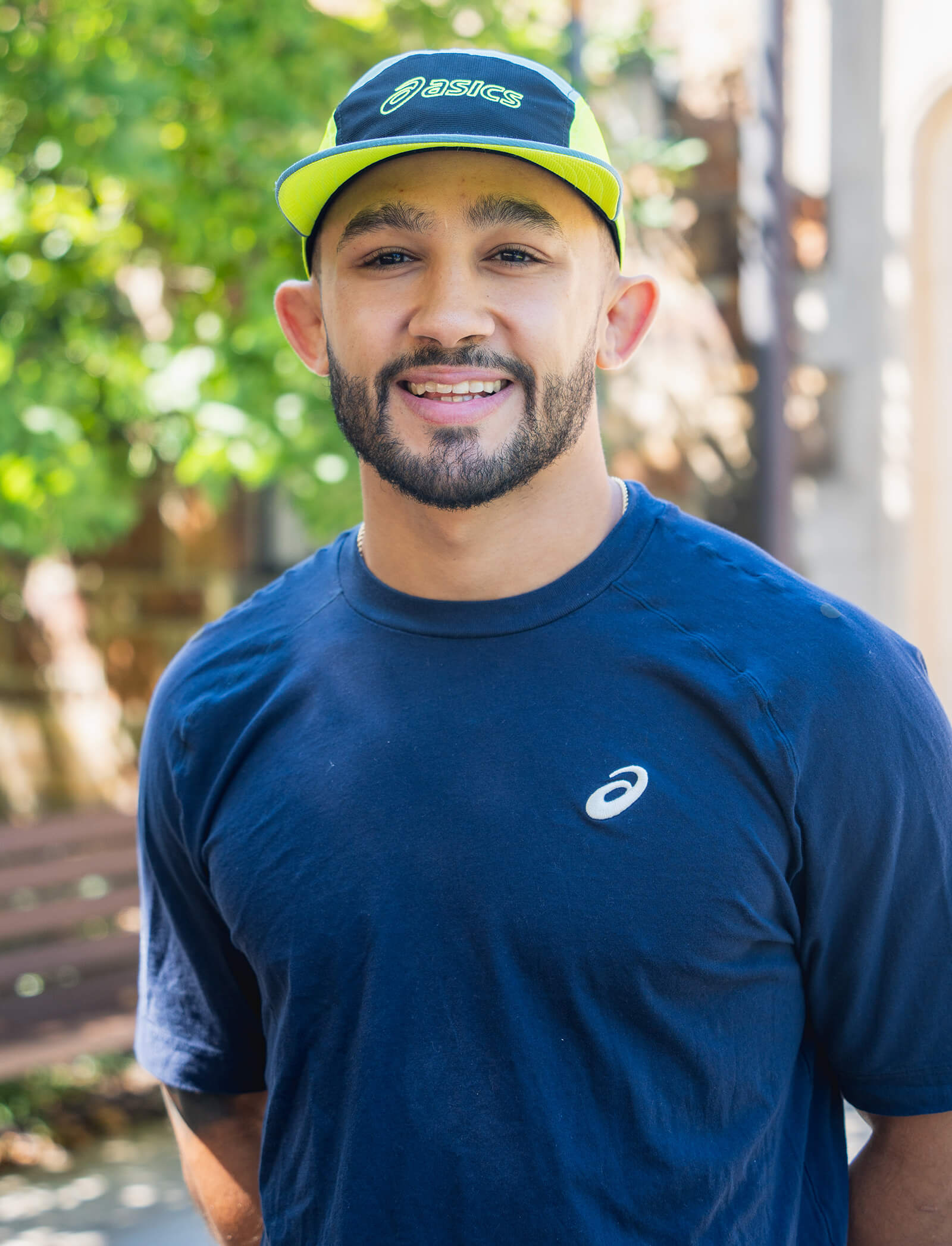 Headshot of Darian Cruz smiling, standing outside, wearing a lime green and navy ASICS hat and navy ASICS tshirt.