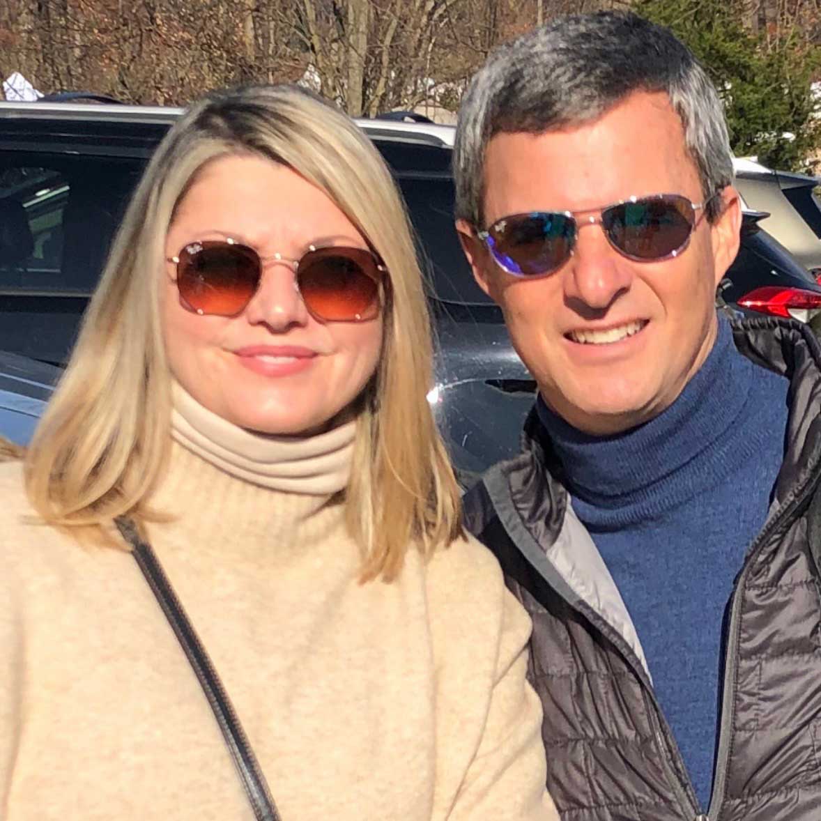 Dave and his wife Mary pose in sunglasses. He wears a dark blue turtleneck and jacket while Mary wears a beige sweater. 