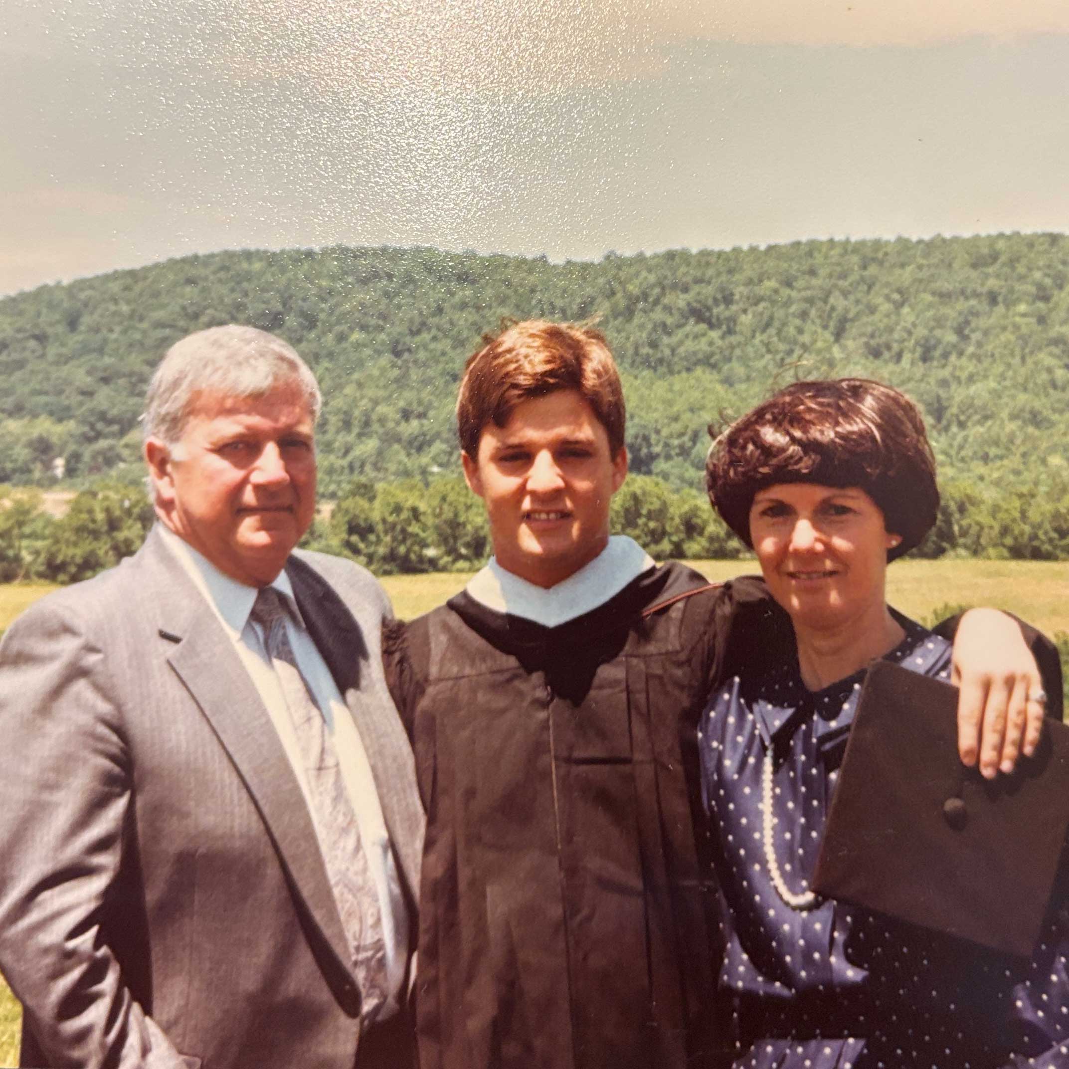 Dave stands outside between his mother and father at graduation. He wears his brown robes while his parents wear business casual. 