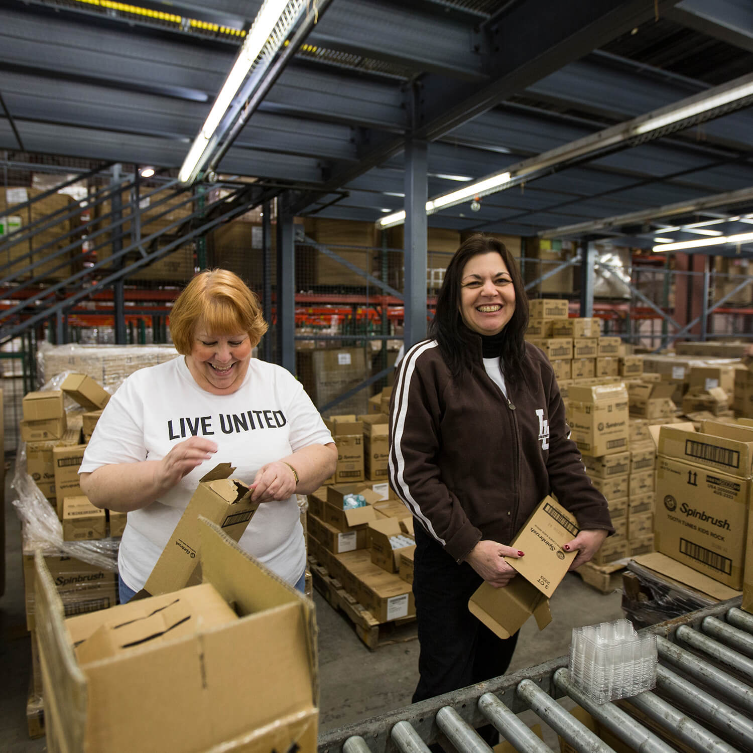 Two women dressed in Lehigh clothing smile as they help with boxing in a warehouse. 