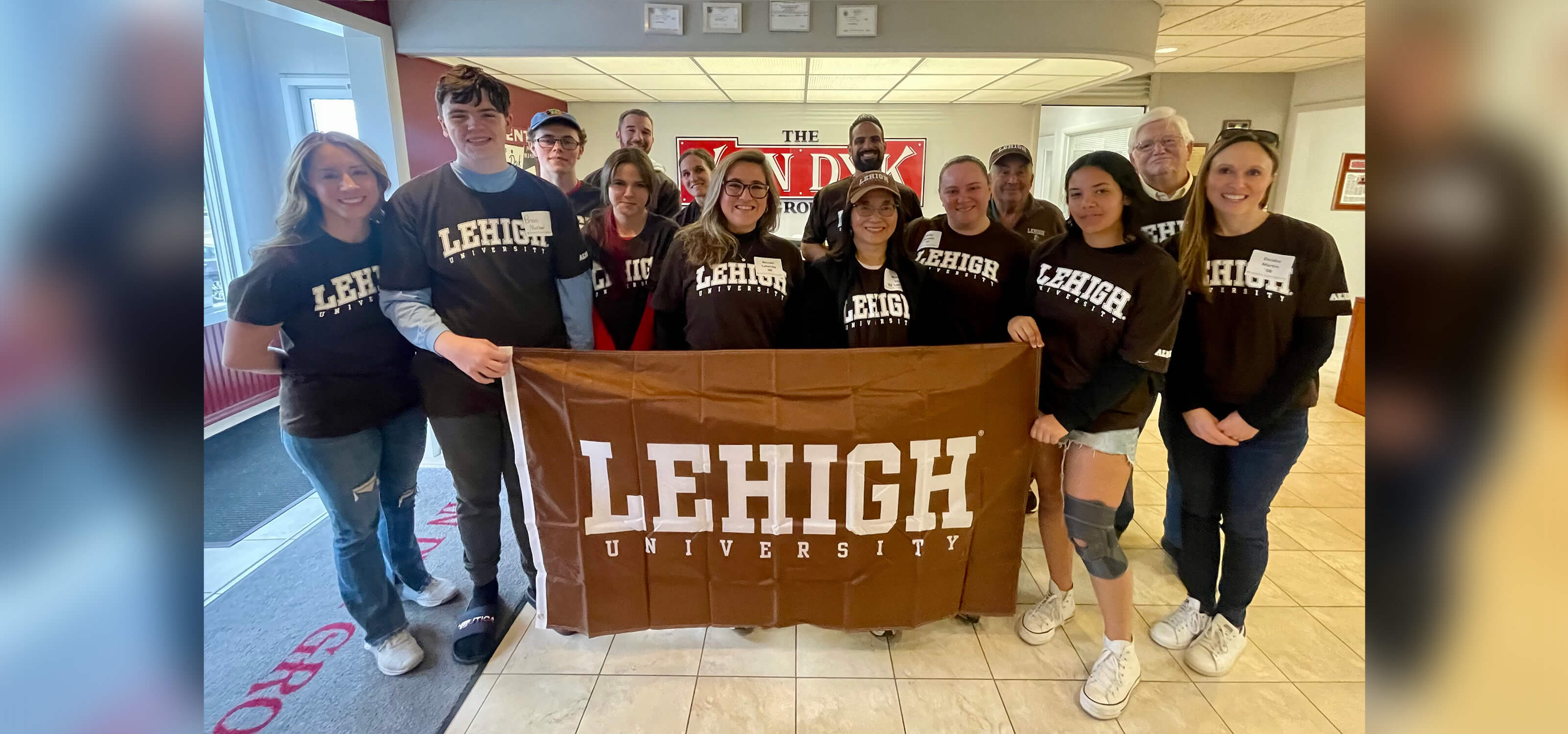 Deidre Martin and her group of alumni volunteers pose wearing brown Lehigh tshirts and holding up a Lehigh flag at the Van Dyk Group headquarters.