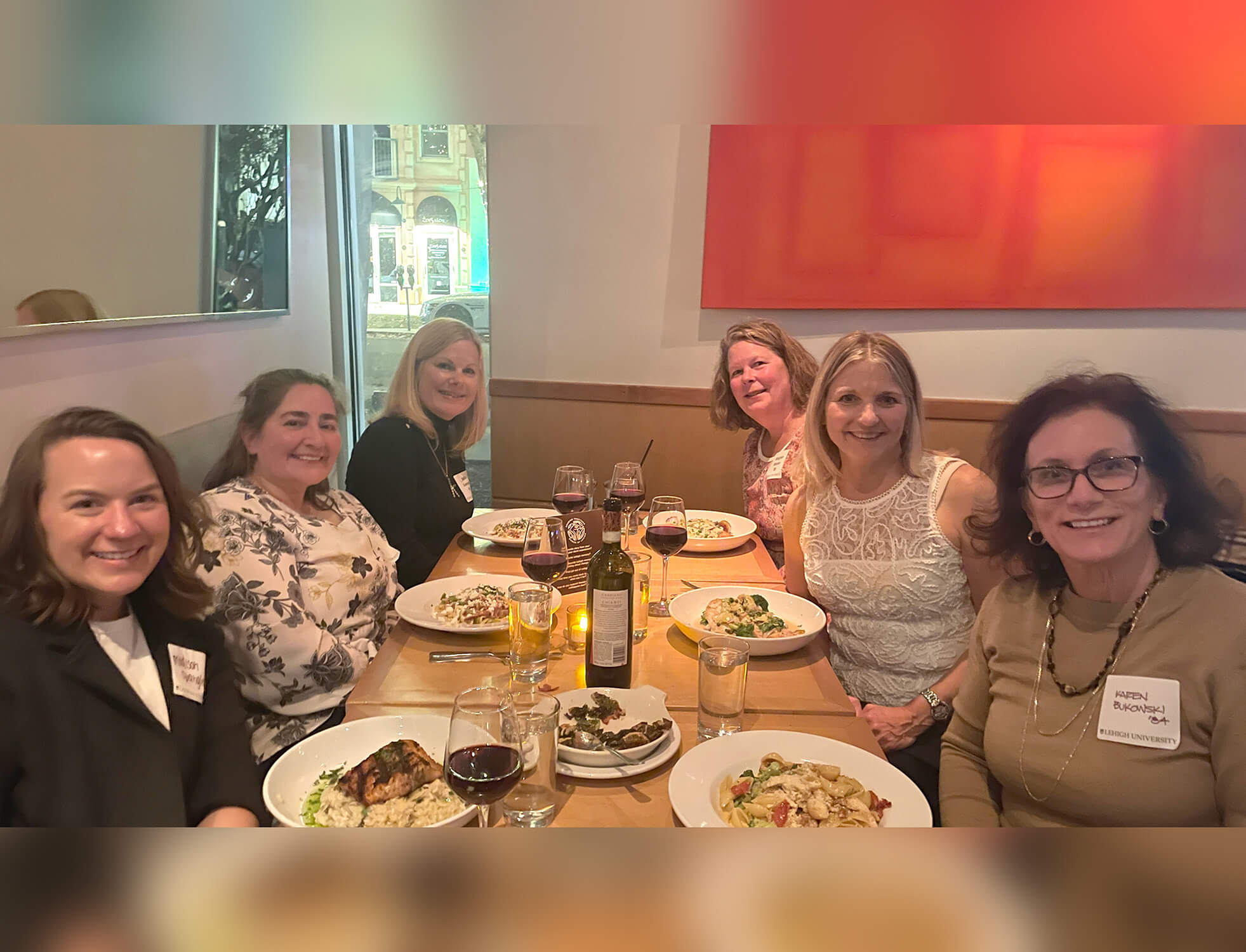 Six women sit at a long table during the day, posing for the photo with full plates of food.