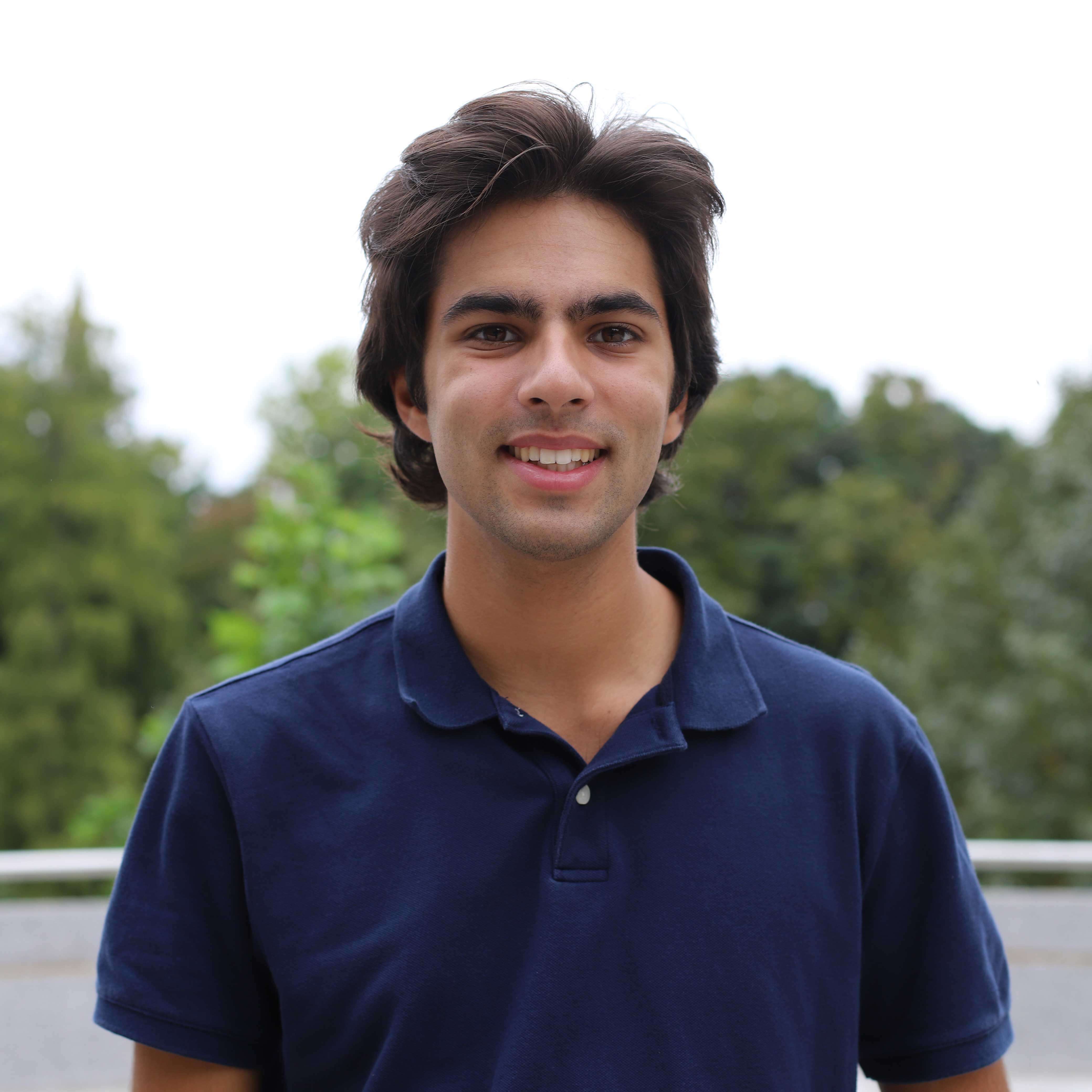Dev wears a deep blue polo shirt and smiles. Trees are visible behind him.