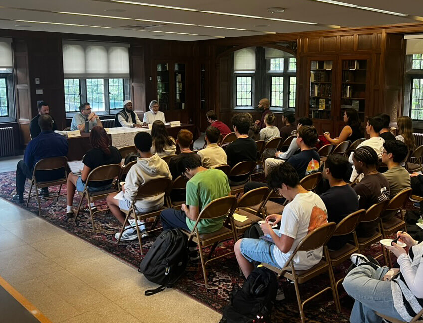 Taken from the back of the room, chairs are arranged for a panel discussion with students in the audience and religious leaders seated at a table in the front.