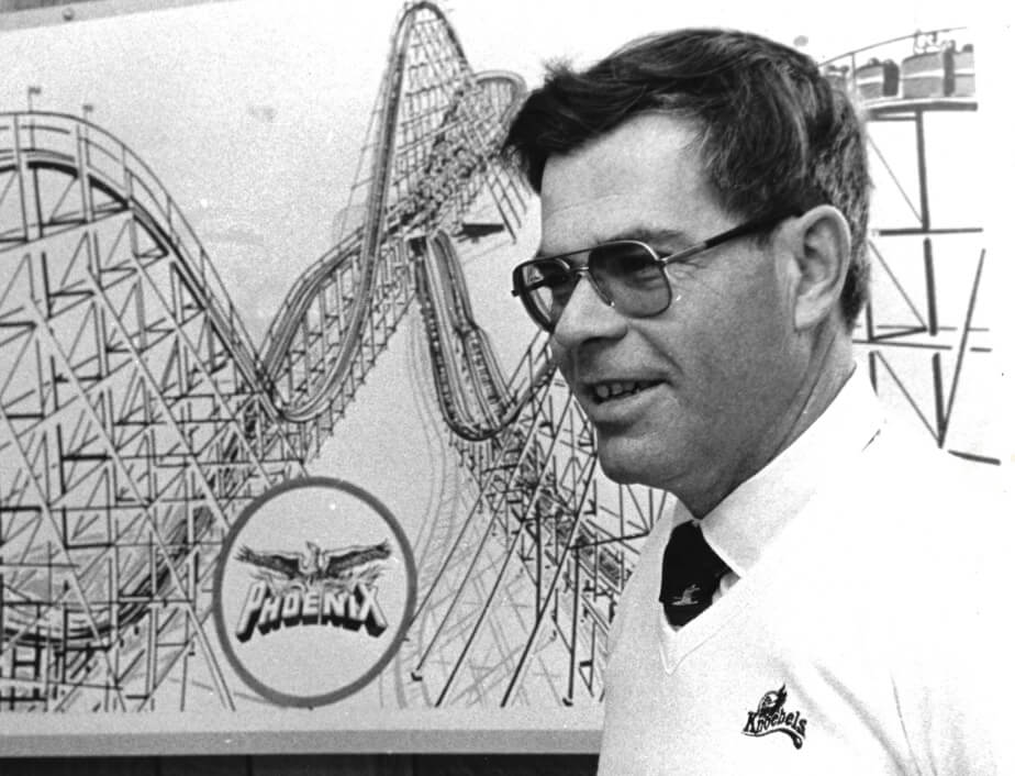 In a black and white photo, Dick Knoebel stands wearing glasses in front of a line drawing of a rollercoaster design for the Phoenix.