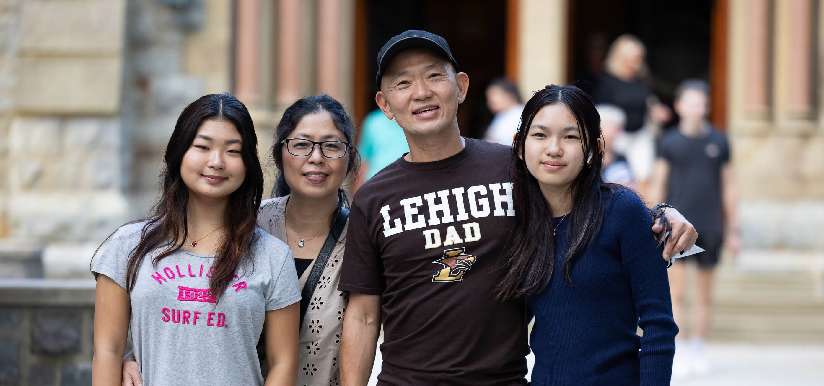 A family of four stands arm-in-arm outside a campus building during Family Weekend with the father wearing a Lehigh Dad shirt.