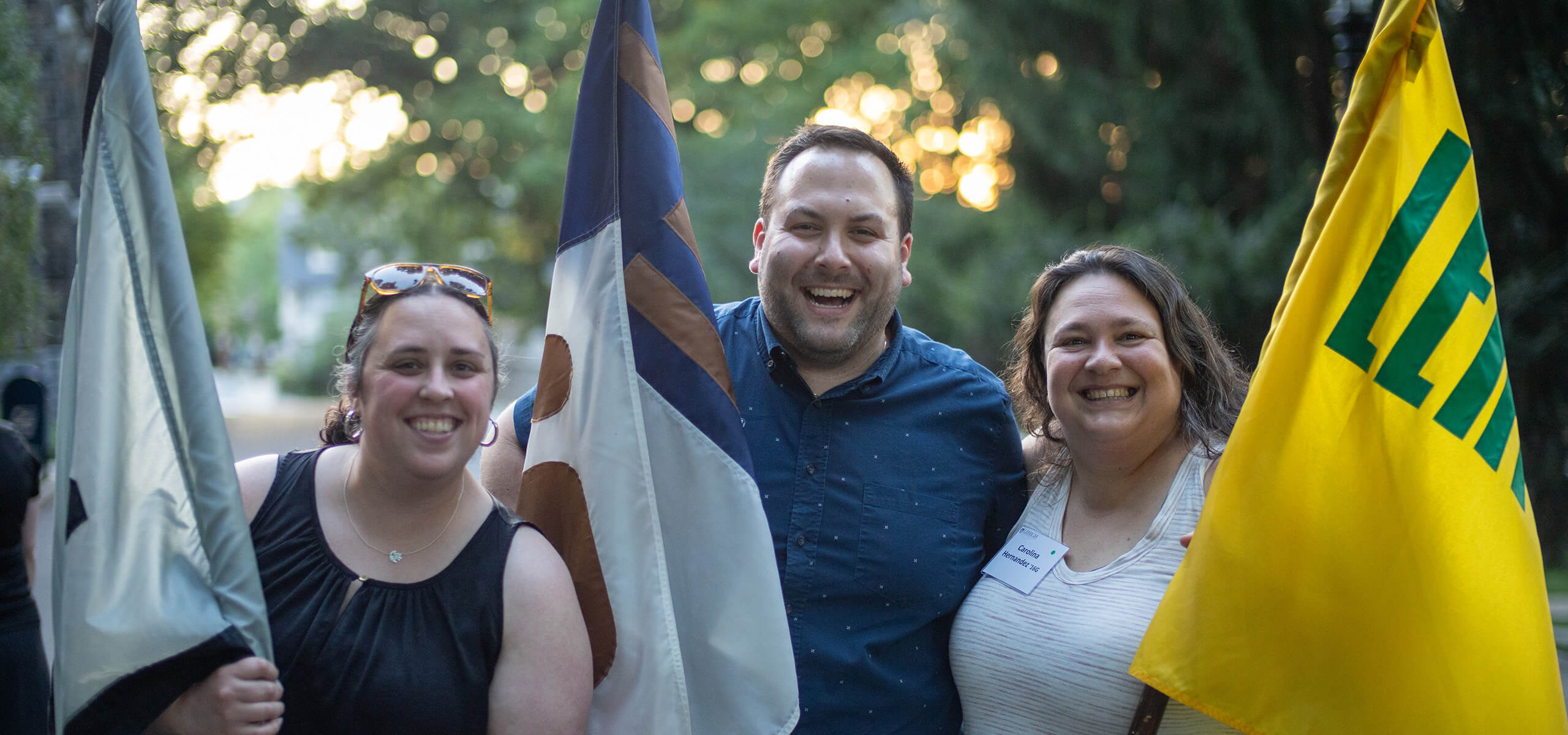 Three alumni stand outside holding their respective class flags with big smiles.