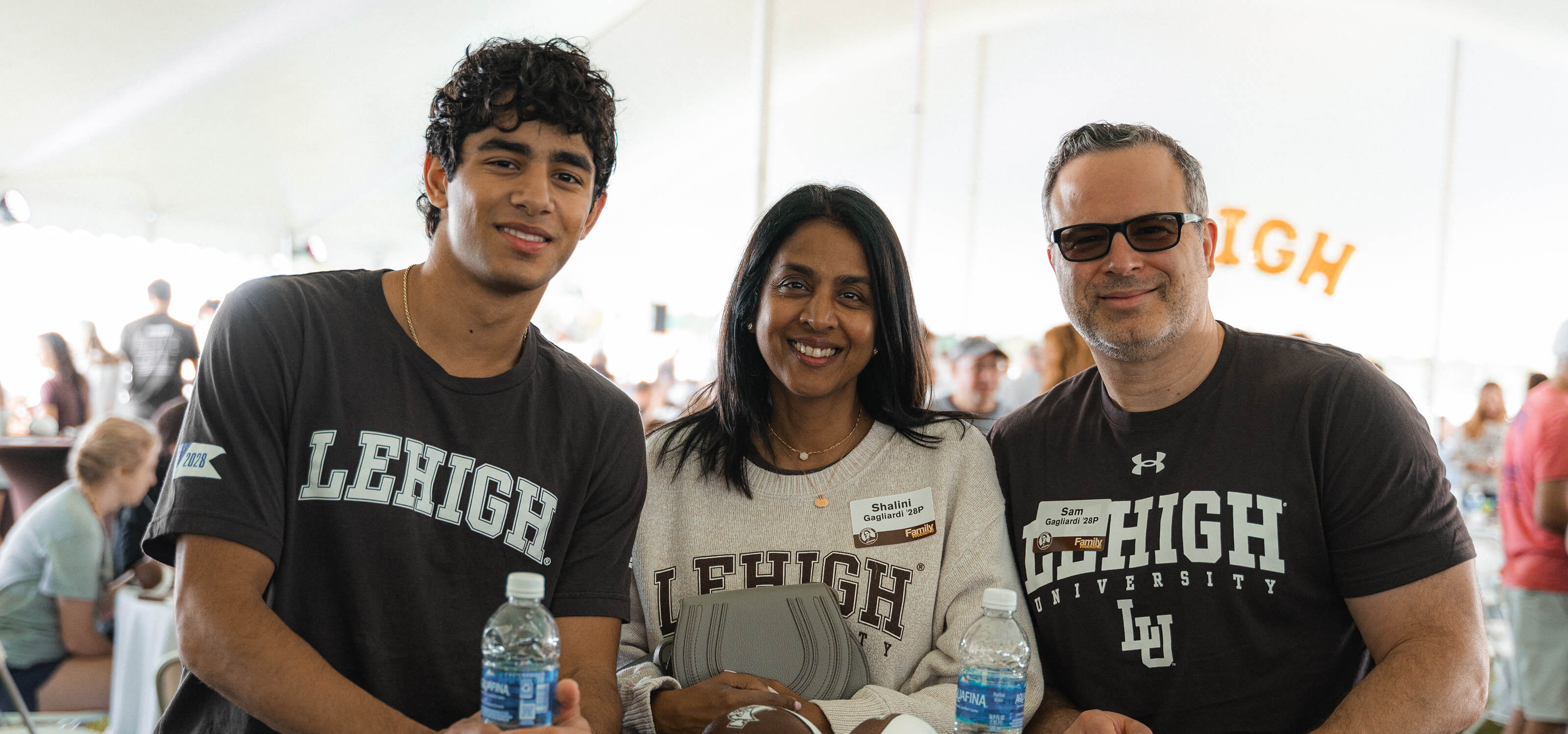 A mother, father and son stand at a hightop table under the alumni tailgate tent during the 2024 Rivalry event, all wearing Lehigh shirts.