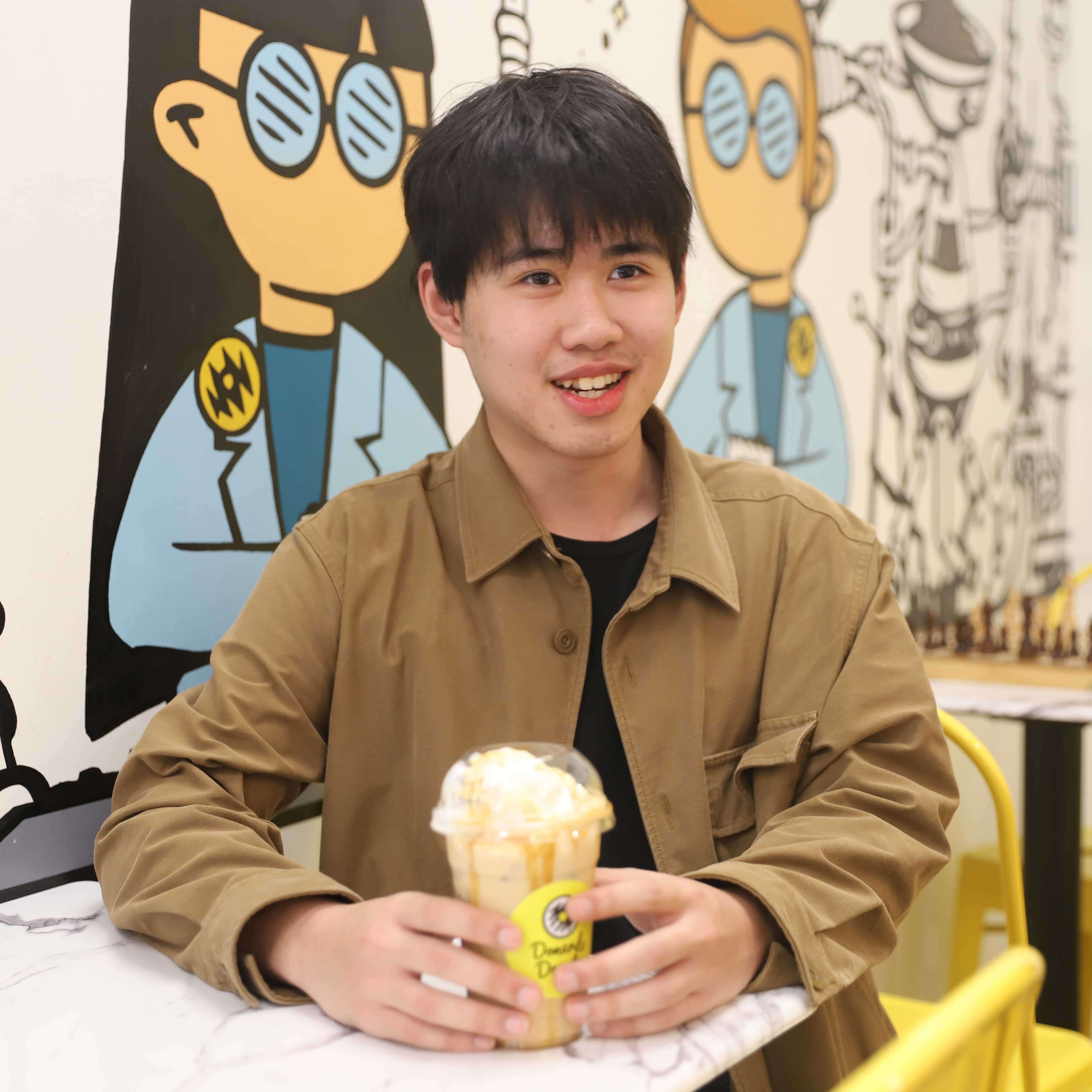 A young man in a brown jacket sits at a marble table with an iced coffee in his hands. 
