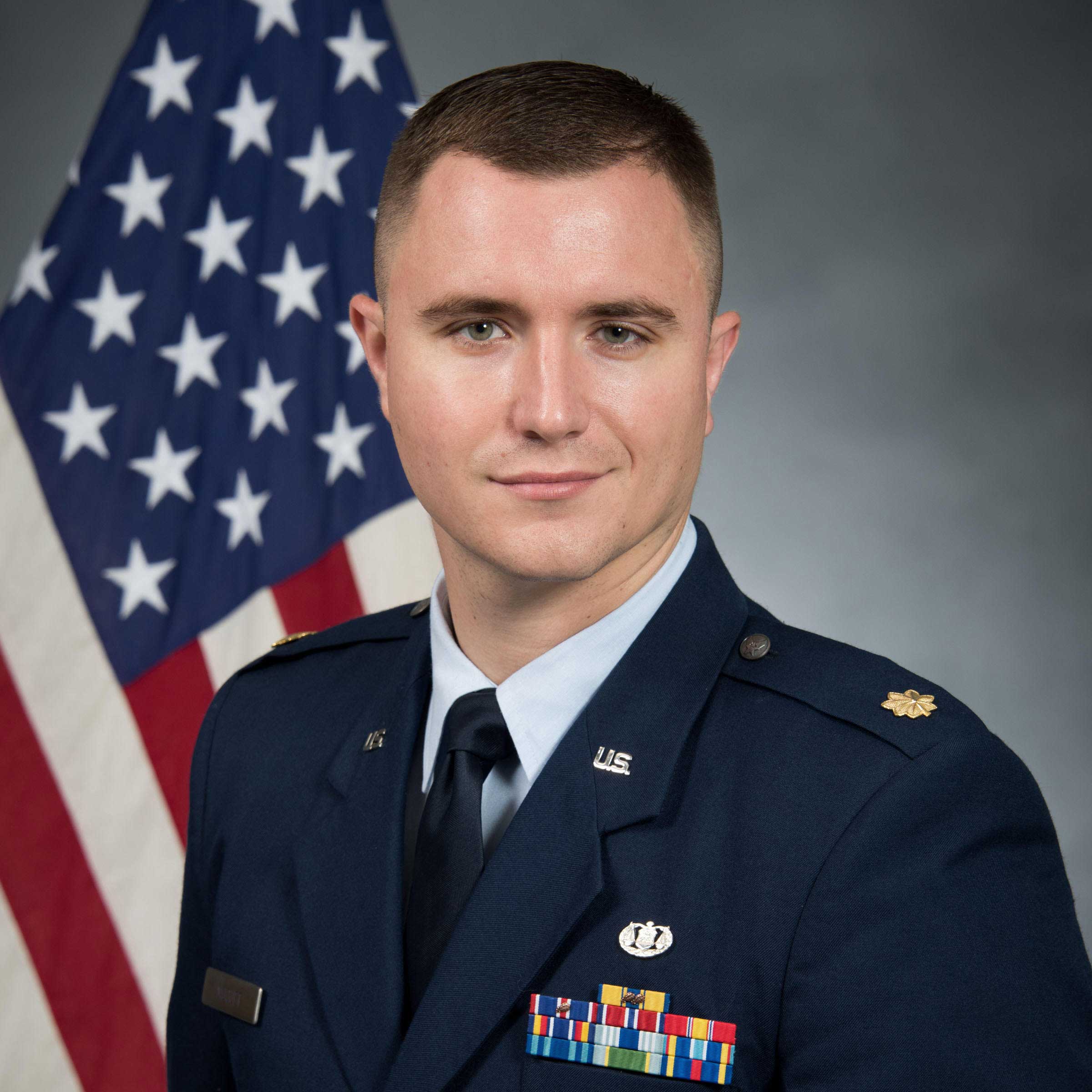 Doug wears a blue suit jacket with several pins decorating it on top of white shirt and tie. An American flag is behind him.