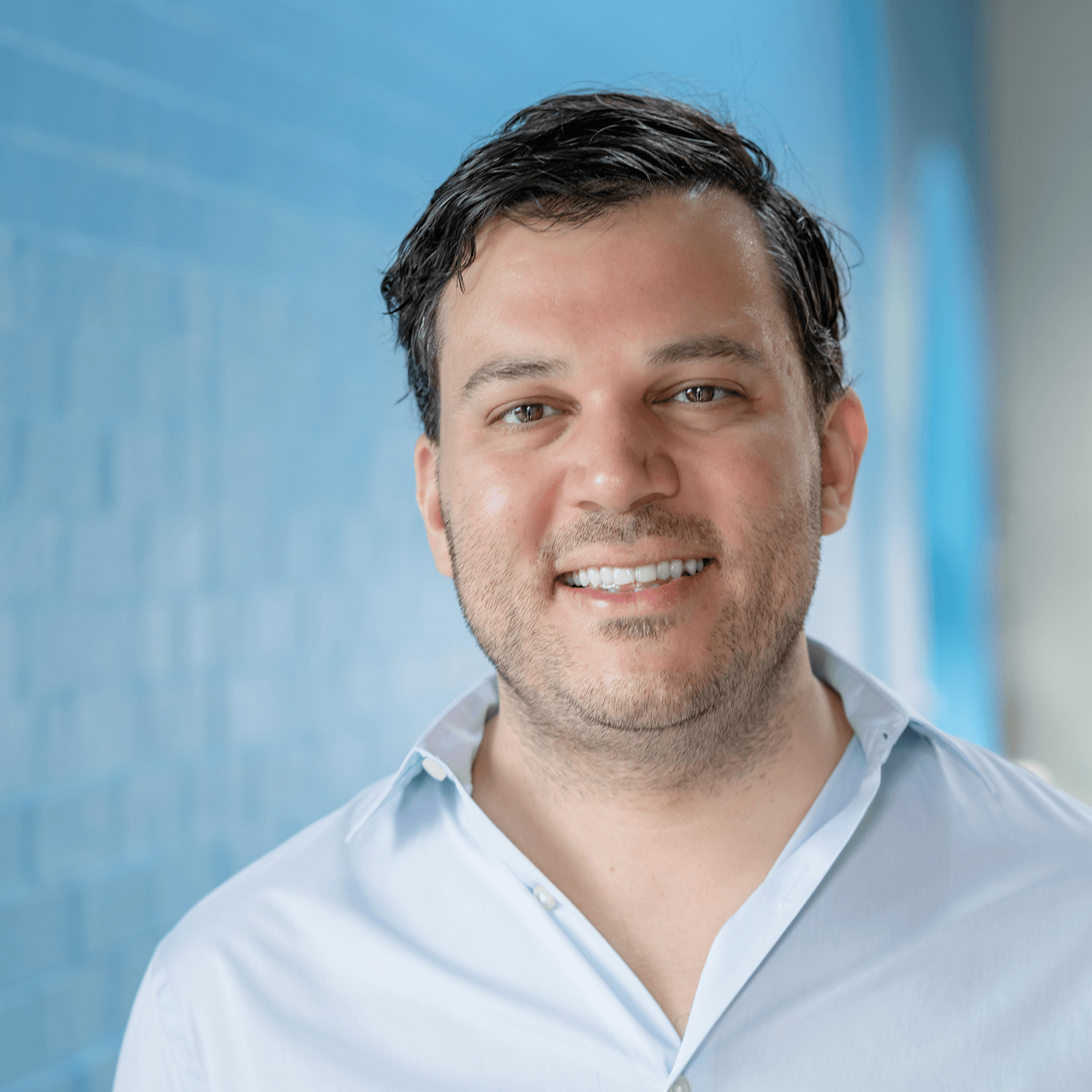 Dr. Amer Alnajar poses for a headshot against a baby blue wall and wearing a light blue business shirt.
