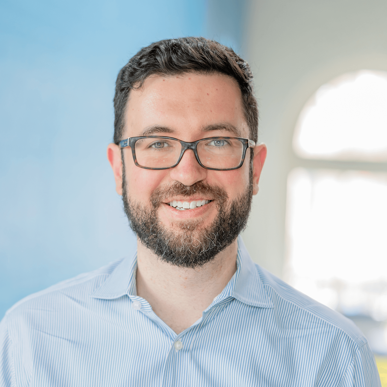 Dr. Hasan Bayat poses with glasses for a headshot in a brightly lit room with a baby blue wall behind him.