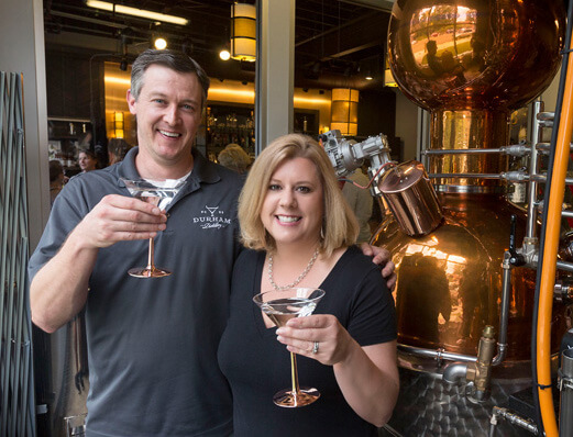 Husband and wife stand side-by-side, in a side hug, each holding up a martini glass in their free hand and smiling at the camera.