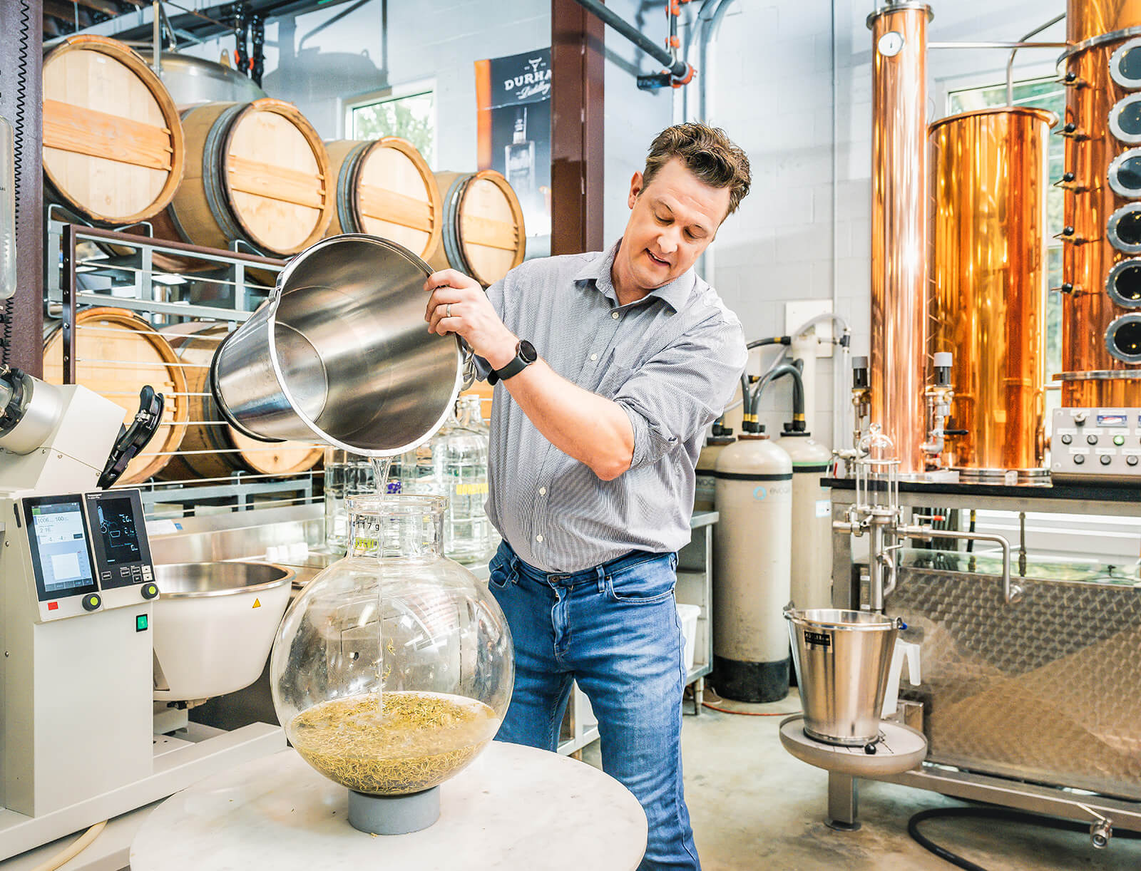 Man stands in a distillery, surrounded by equipment and barrels, carefully pouring ingredients.