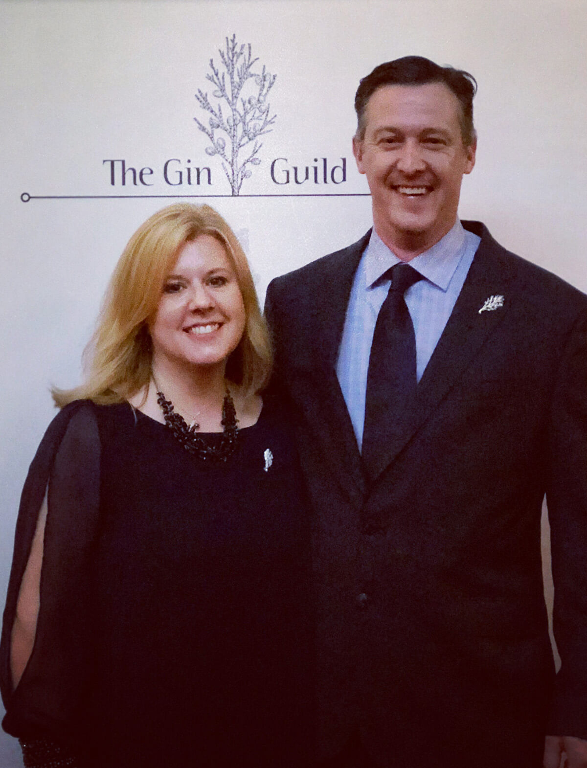 Husband and wife stand side-by-side dressed elegantly and standing in front of a sign for the Gin Guild, smiling at the camera.