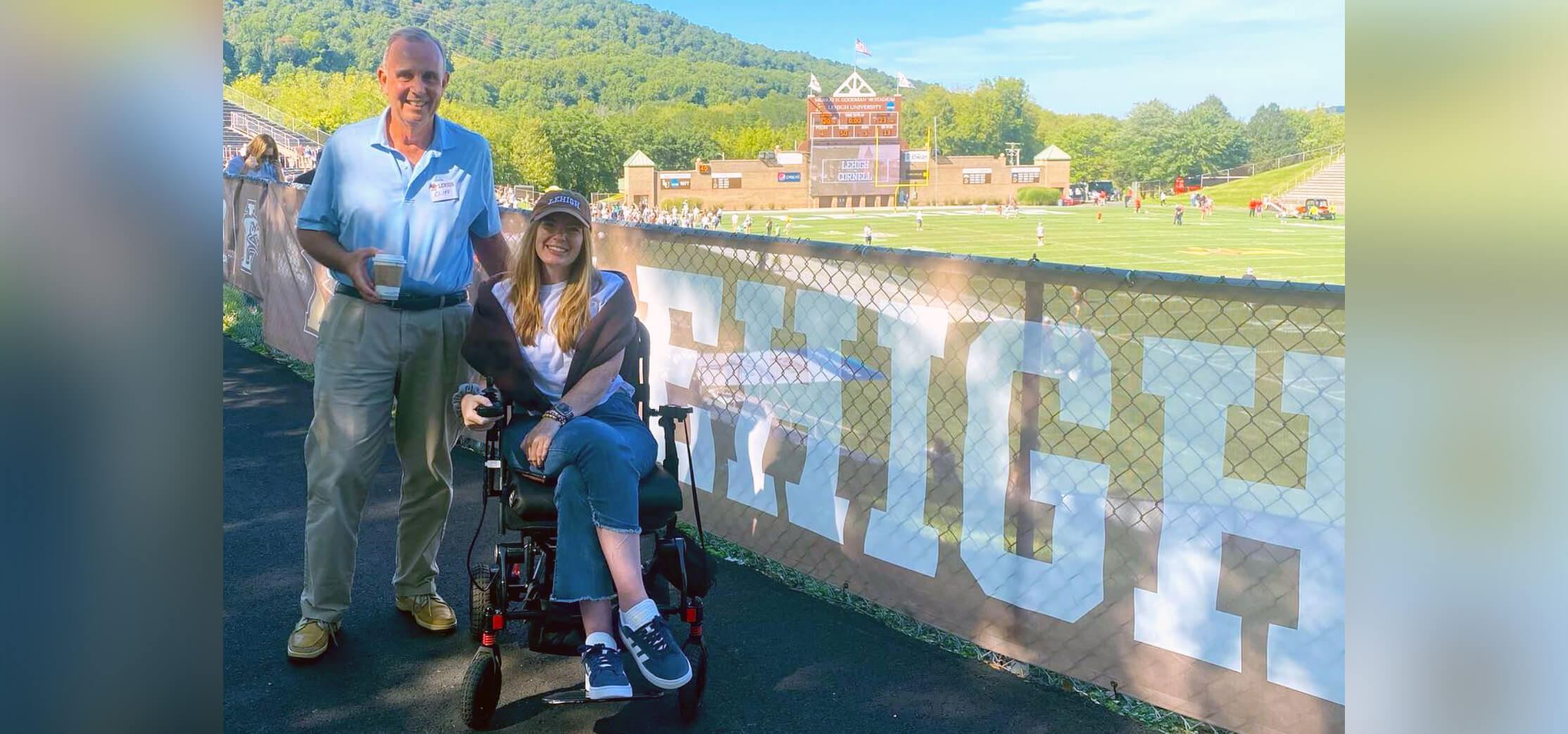 Brooke Eby seated wearing a brown Lehigh cap in front of a Lehigh banner hung on a fence overlooking Goodman Stadium, her father standing beside her.