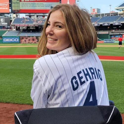 Seated in her chair, BrookeEby turns to look back at the camera over her shoulder, showing a pin-striped jersey with “Gehrig” on the back.