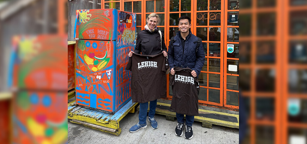 Two Lehigh alumni hold up brown Lehigh tshirts in front of a colorful storefront with a refrigerator pained in vibrant colors.