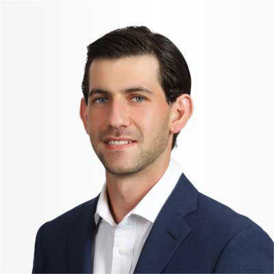 Elan wears a navy suit jacket with a white button up underneath. He stands in front of a white background and smiles.