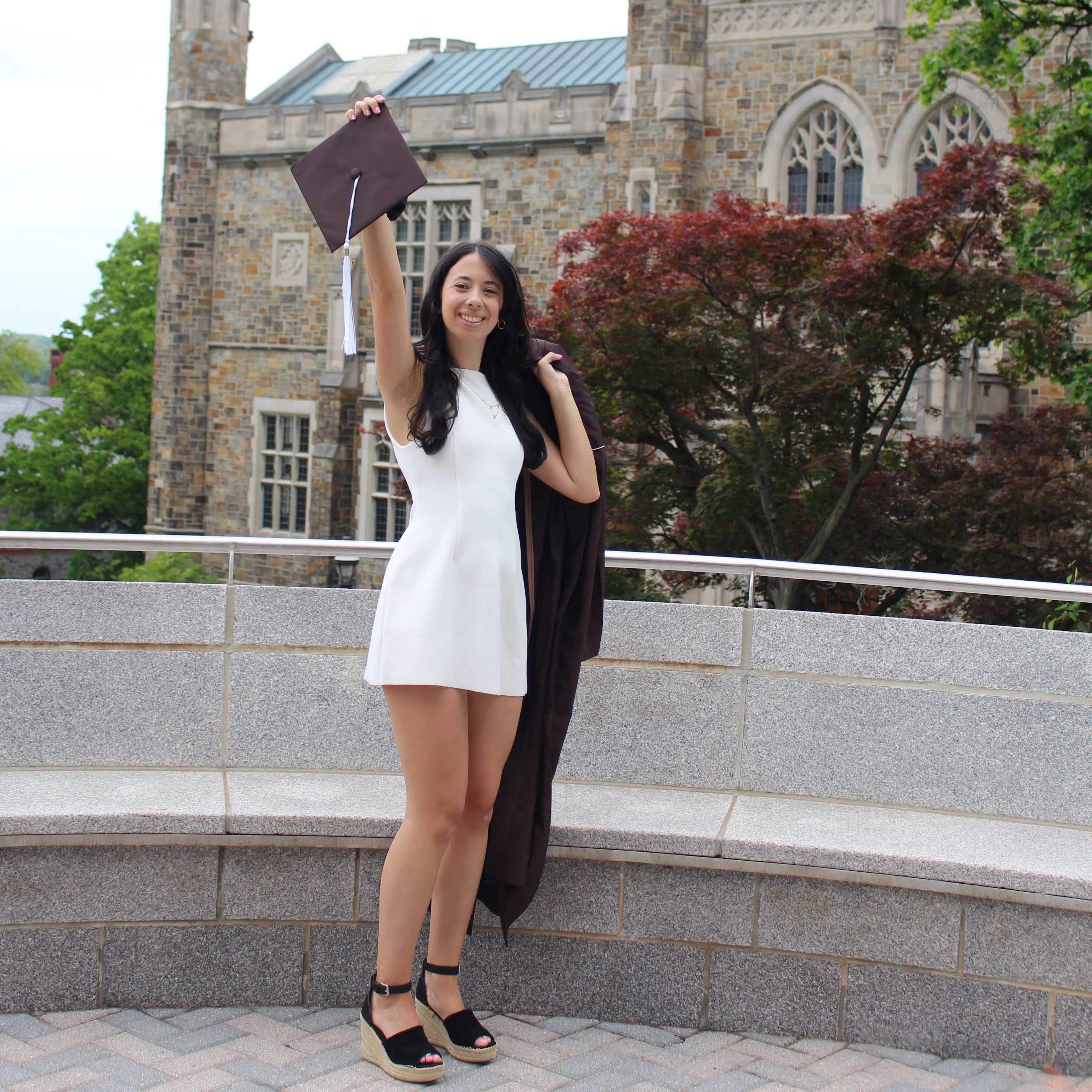 Emily wears a white dress and holds her graduation gown. She extends her arm and holds up her cap.