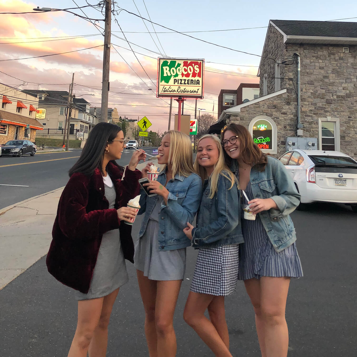 Four college aged women hold Rita’s Water Ice outside a stone building a “Rocco’s Pizzaria” sign as one feeds another from her spoon.