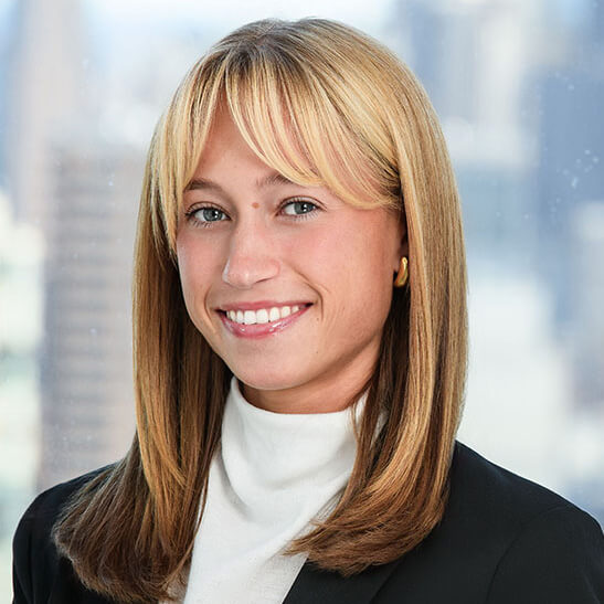 Emily Segal smiles in a dark suit jacket and white mock turtleneck, wearing her blonde hair straight and shoulder length.