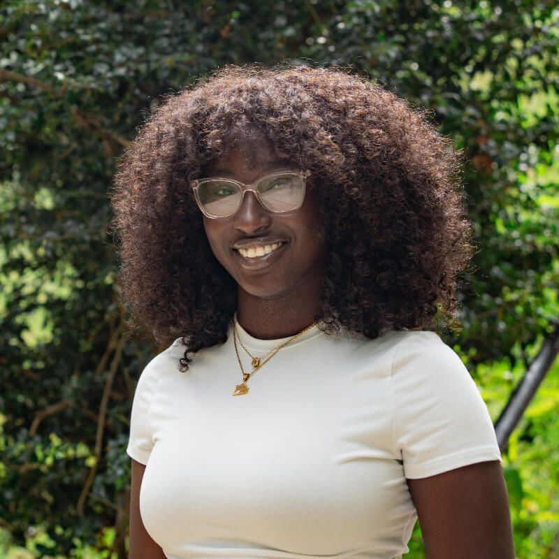 Emmanuella Agyemang poses outdoors for a headshot, with green foliage in the background, wearing glasses, a white shirt, and layered gold necklaces.