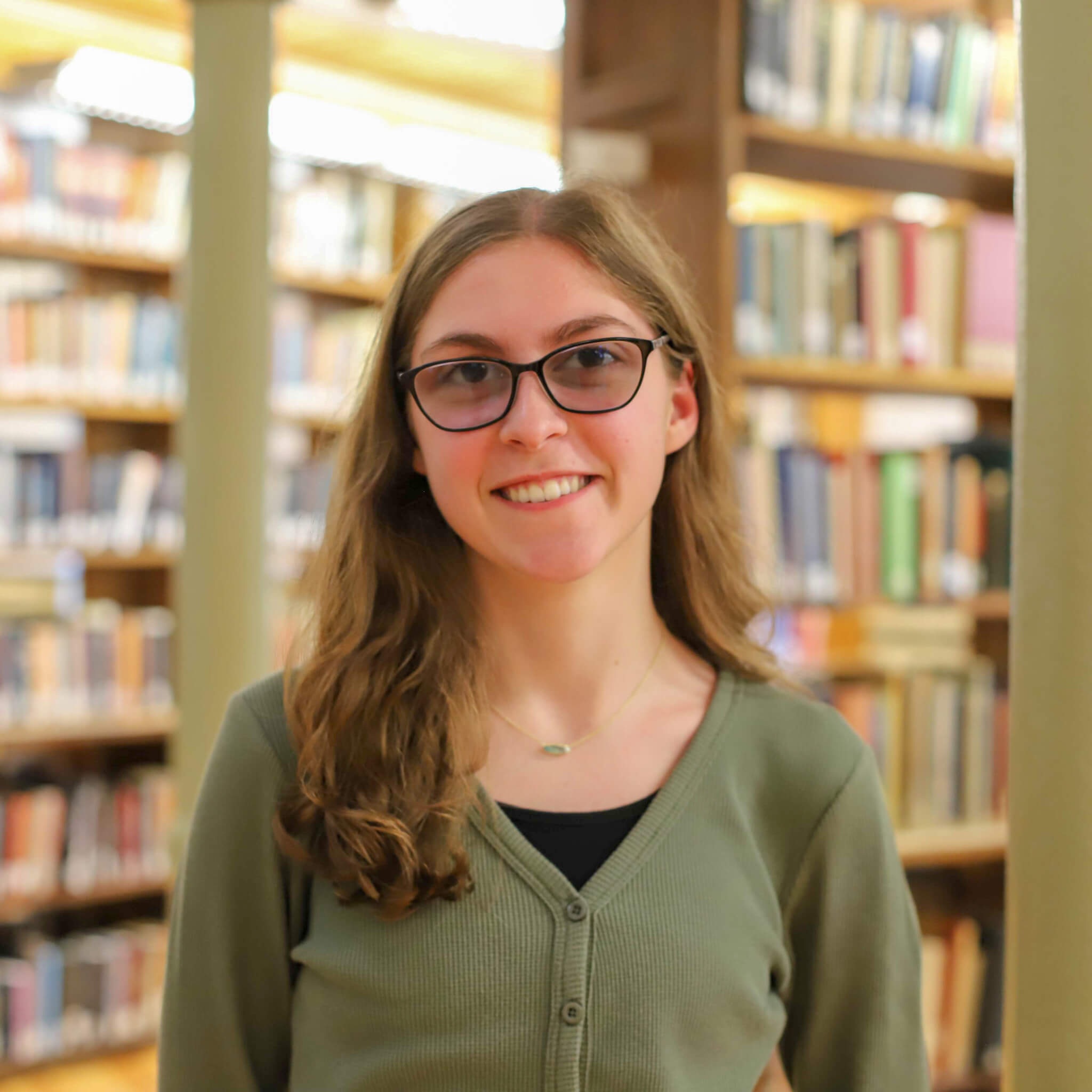 Erica wears a black camisole with a green cardigan atop and black glasses. Shelves of books are visible behind her.