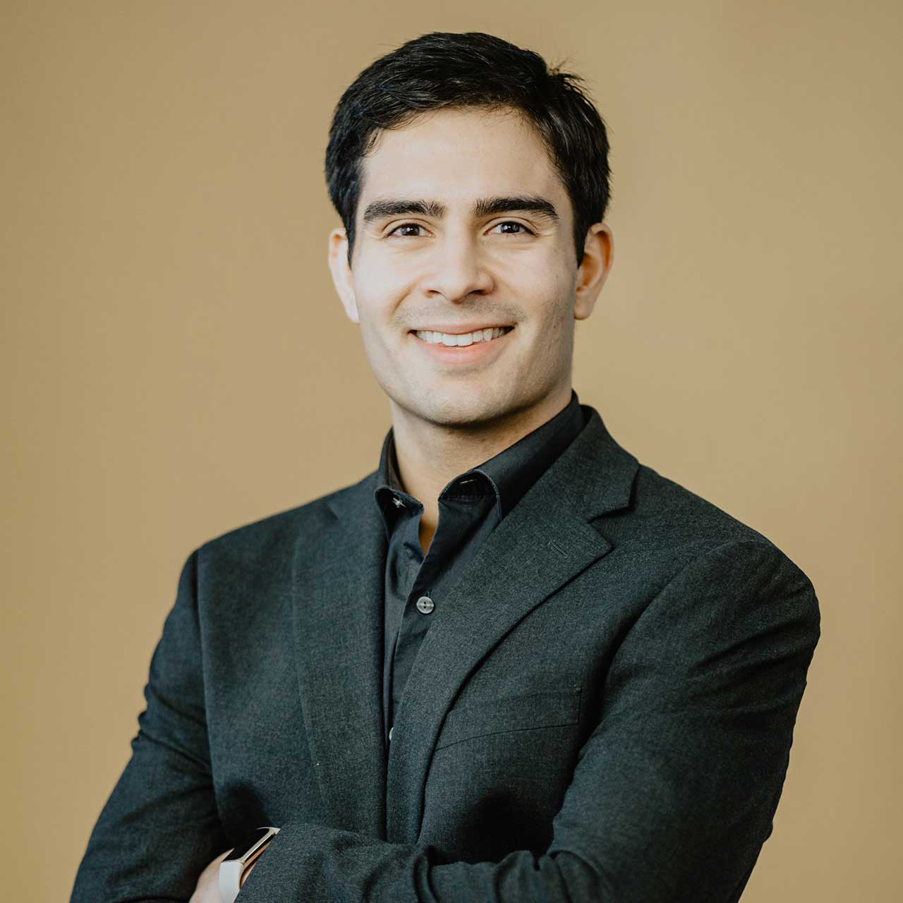 Erik wears a black suit jacket with a black button up underneath. He stands in front of a beige background and smiles.