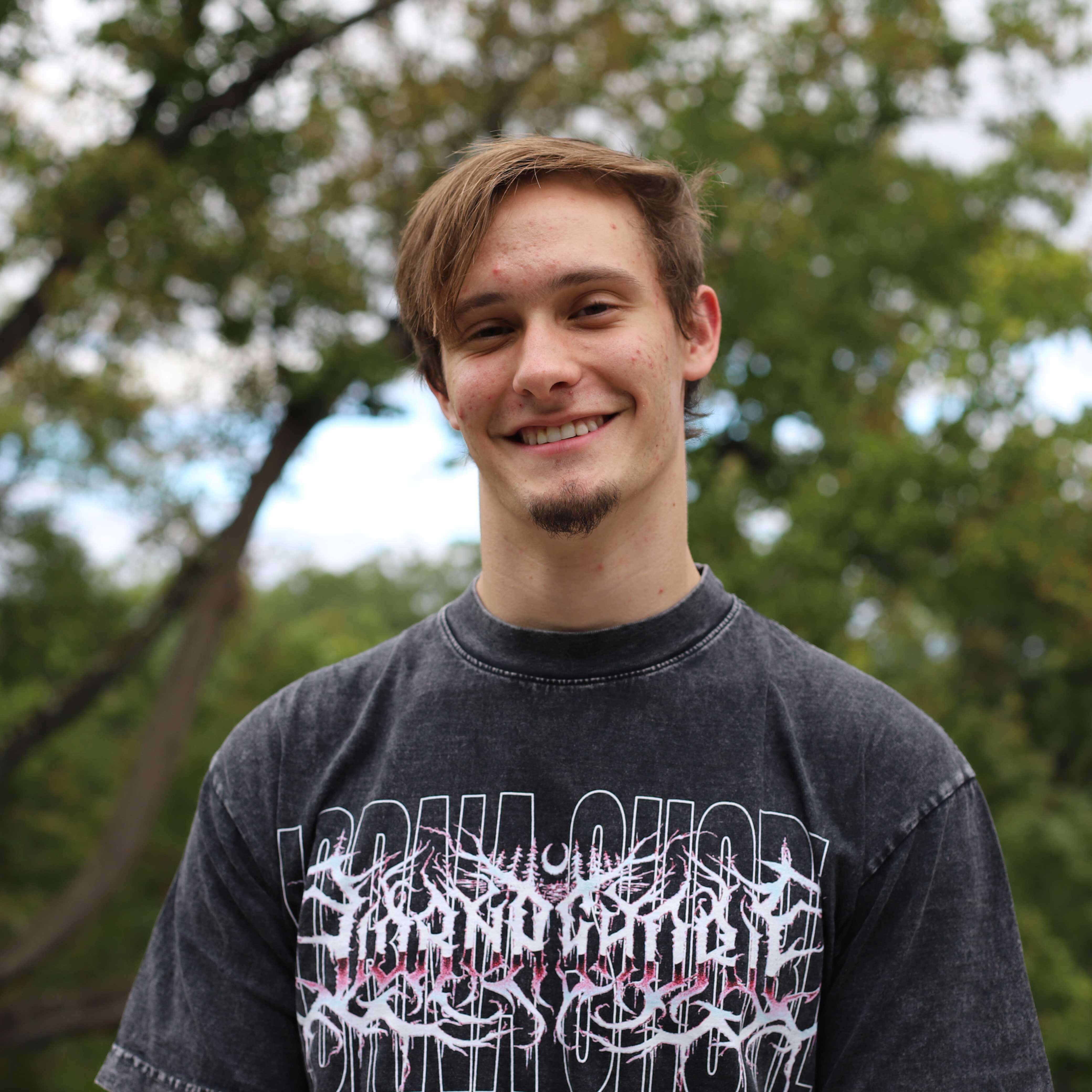 Evan wears a gray, acid-washed t-shirt and smiles. Trees are visible behind him.
