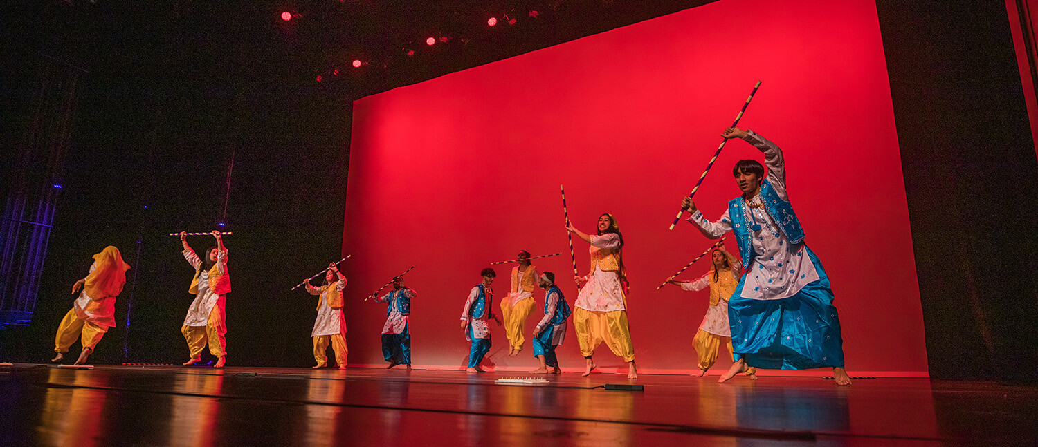 A group of dancers on stage perform energetically against a vibrant red backdrop, holding colorful sticks.