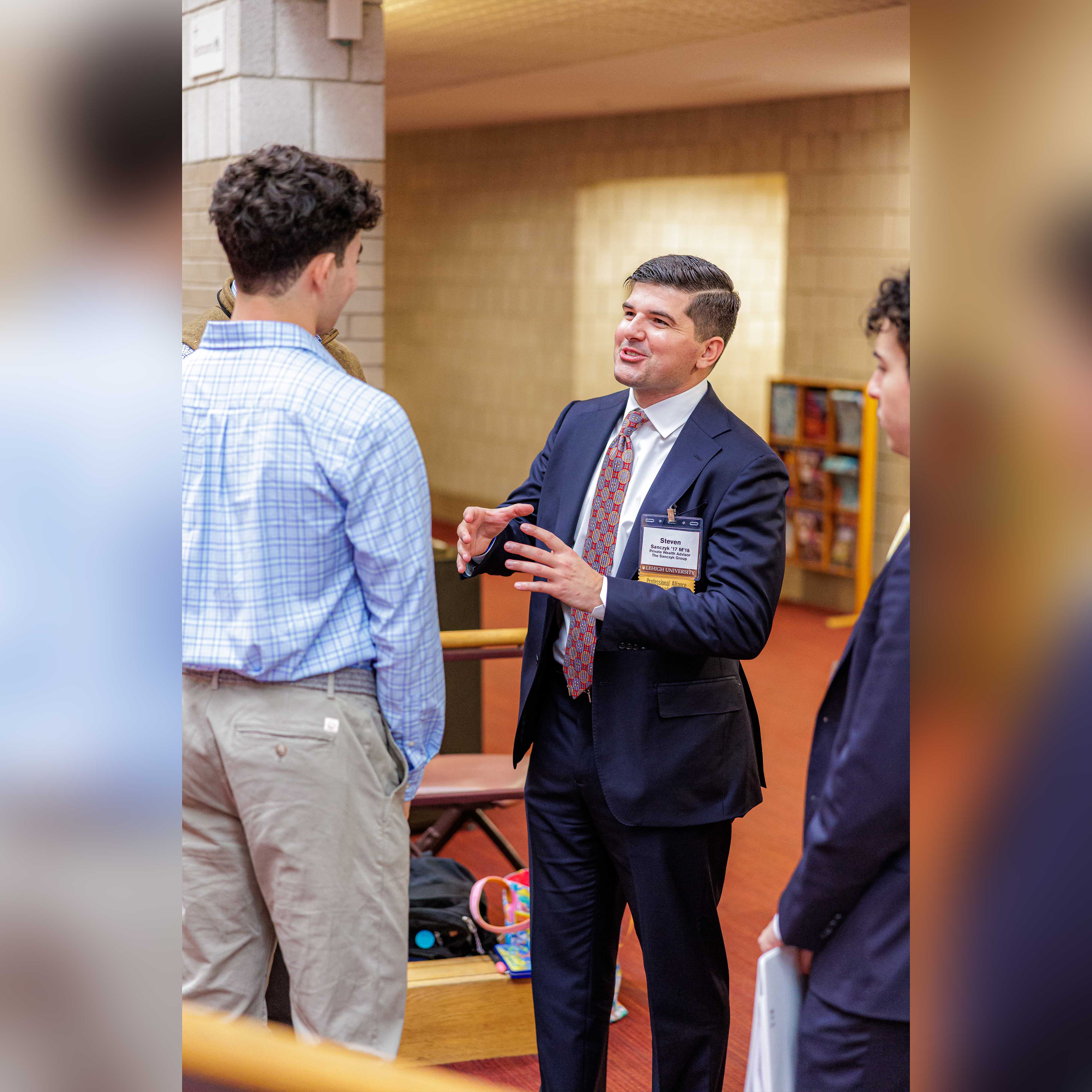 An alum in a navy suit and red tie smiles while talking to attendees that also wear business casual attire.