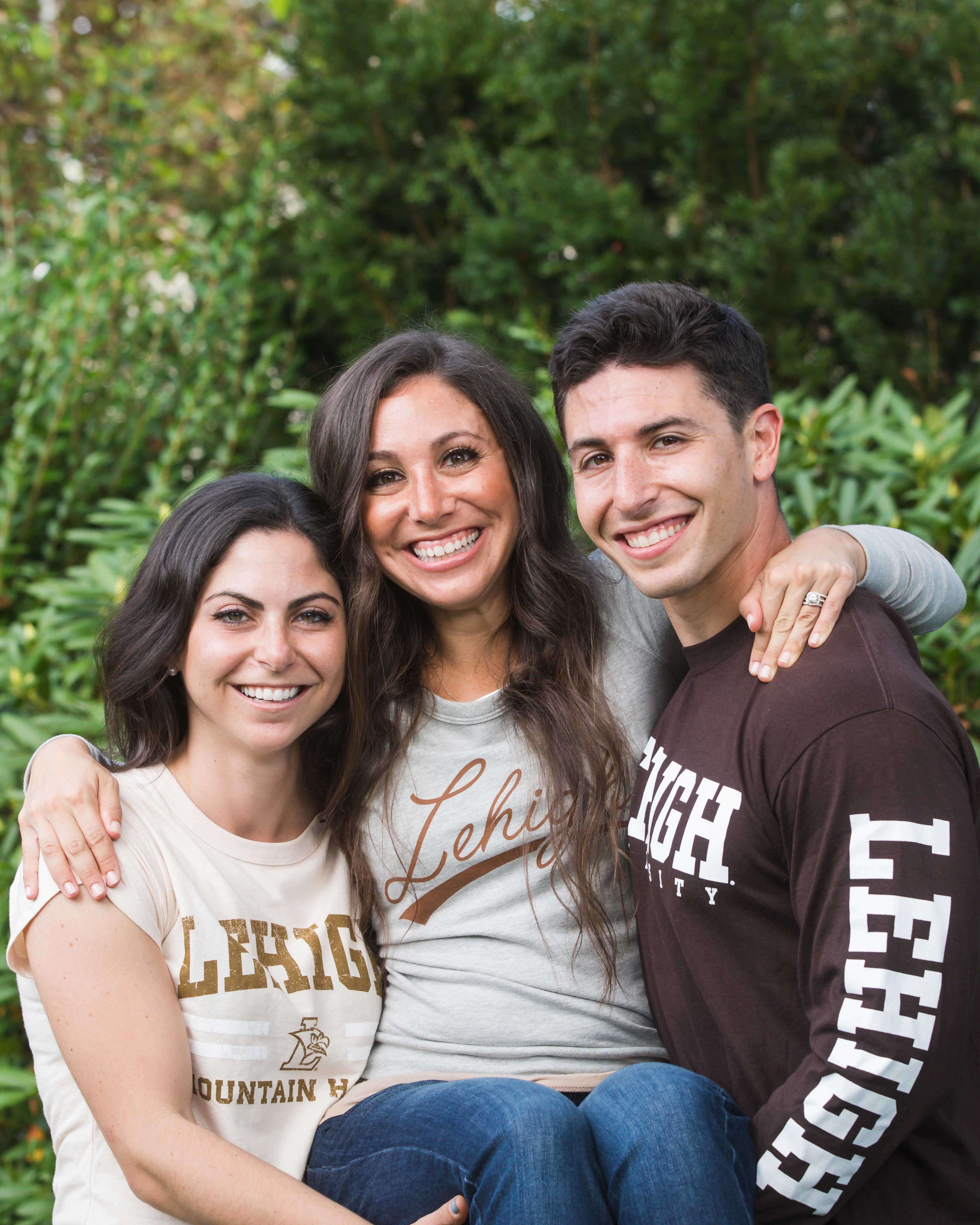 Allison and Max hold Jessica and smile. They each wear Lehigh-branded tops in neutral shades. The light is warm; greenery nearby.