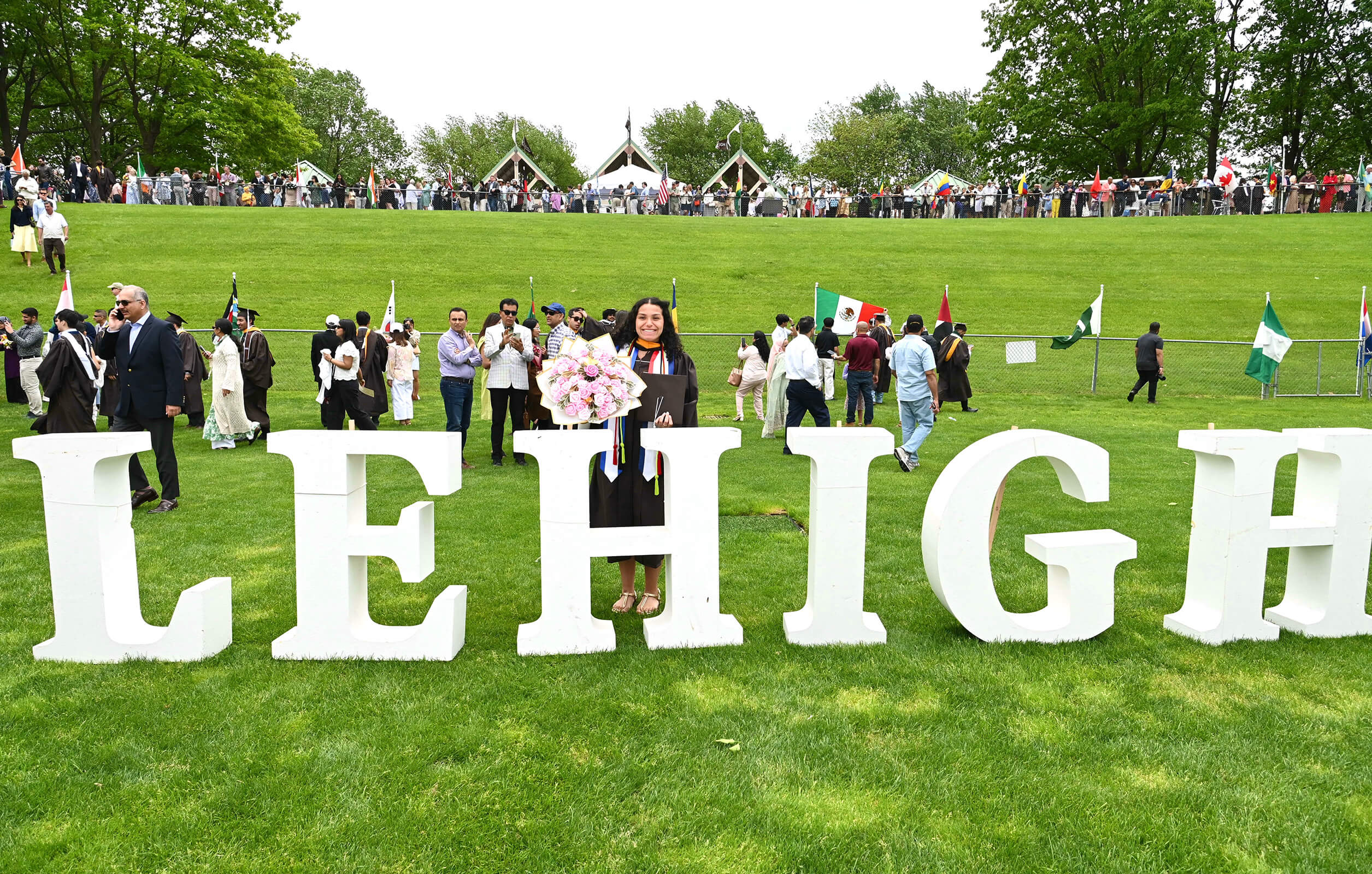 Arianna Morataya stands behind large letters reading “Lehigh” on a green lawn on commencement day, wearing her brown robes and holding a large bouquet of flowers. 