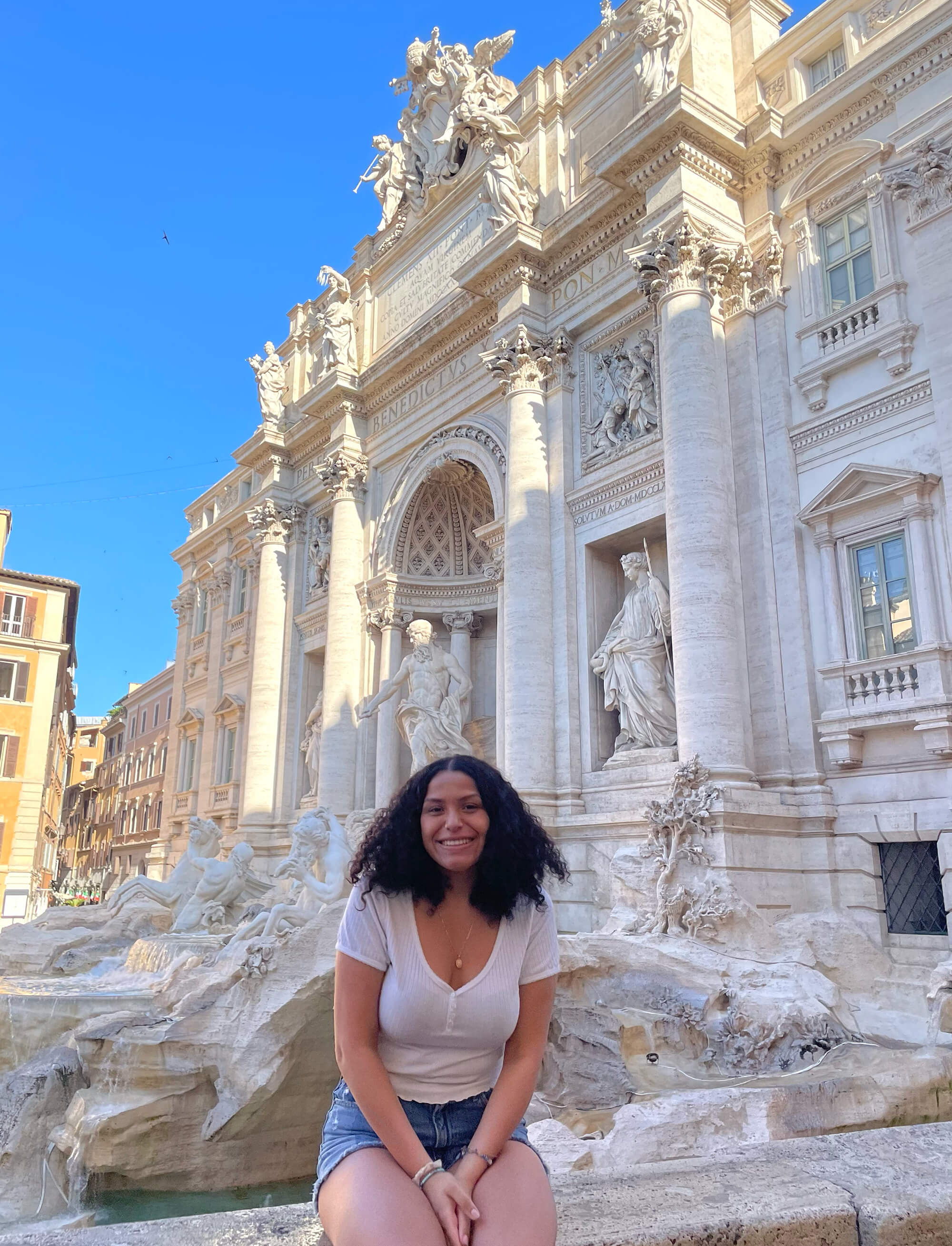 Arianna Morataya sits on a stone wall in front of the Trevi Fountain in Milan, wearing a tshirt and shorts on a clear day.