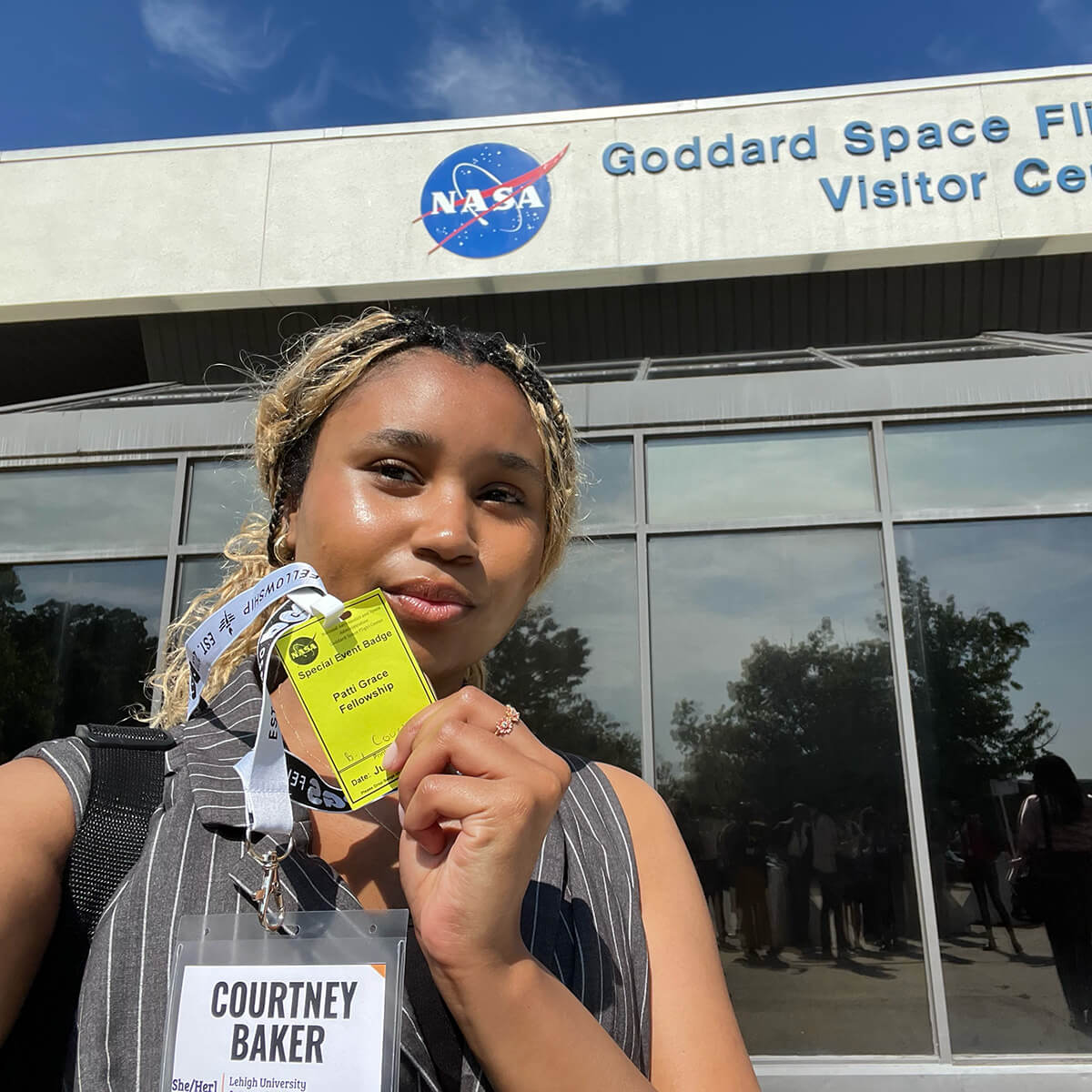Courtney Baker stands in front of a NASA building and holds a yellow event badge that lists the name of the fellowship. 