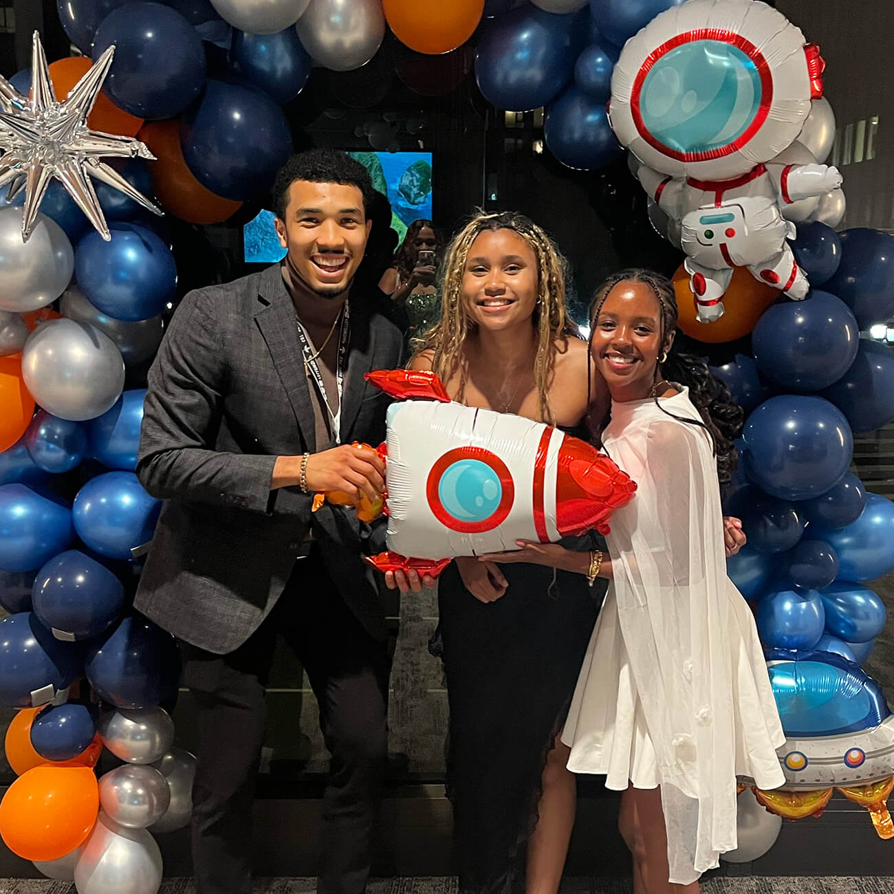 Courtney Baker, Alyssa Weeks Jenkins, and Harrison Jenkins stand under a space-themed balloon arch holding a rocket balloon.