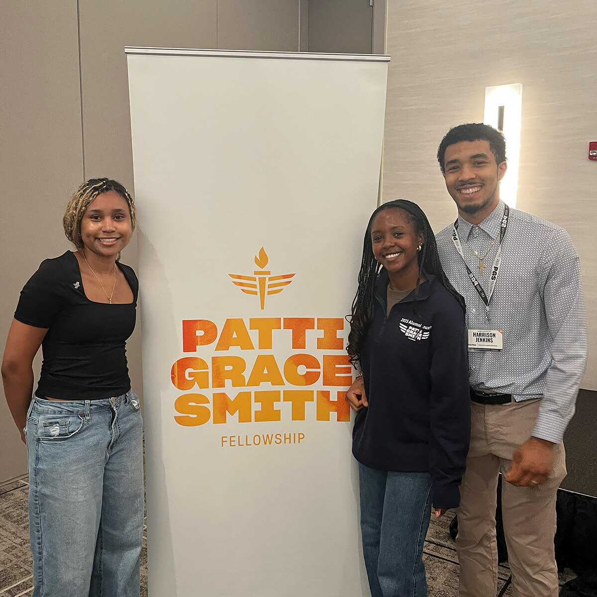 Courtney Baker, Alyssa Weeks Jenkins, and Harrison Jenkins stand near a fellowship banner; a silver pin is on Courtney’s top.