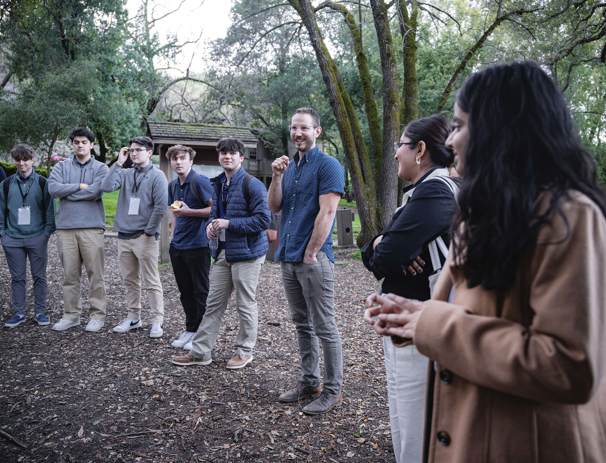 A man stands in the center of a row of students, speaking outside on ground covered in brown leaves and dirt.