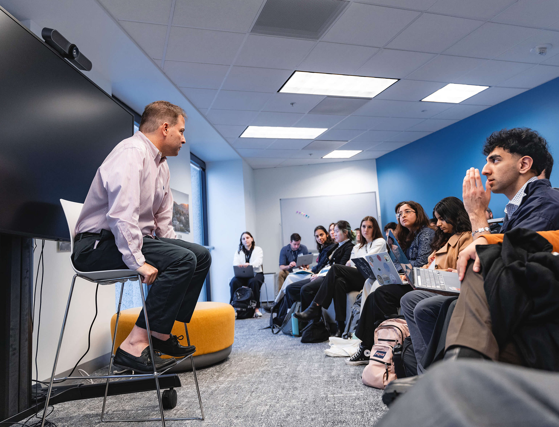 A man sits on a counter height chair in front of a group of students with laptops on their laps.